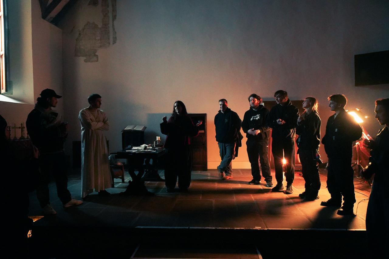A group of eight people stands in a dimly lit space. One person in a white robe gestures while speaking, surrounded by others in casual clothing, including hoodies and jackets. A table with items is positioned in front of them.