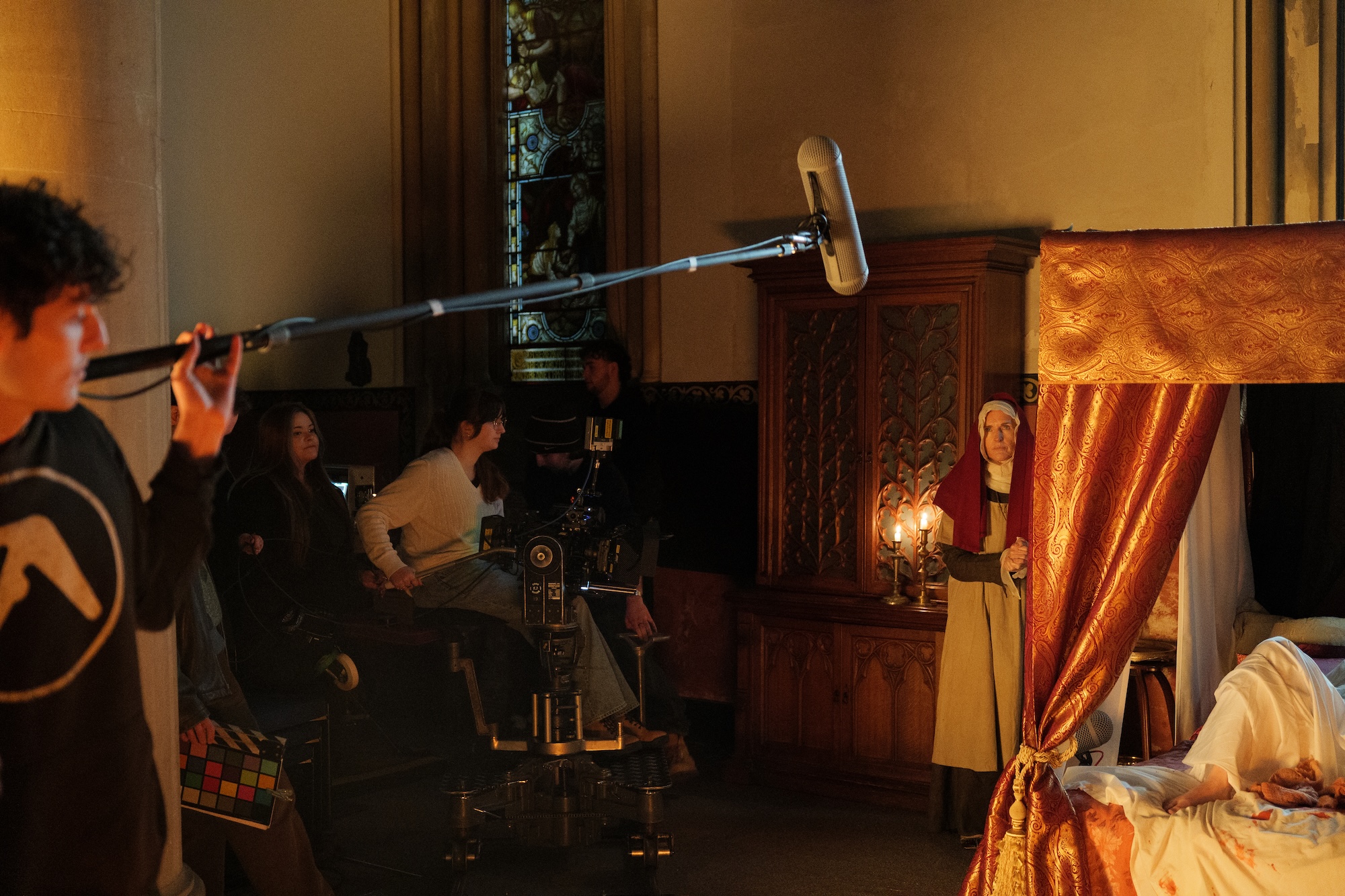 A sound technician holds a boom microphone above a scene set in a historic room. Actors are positioned near a large bed and a wooden cabinet while a person sits behind a camera on wheels. Candlelight illuminates the space.