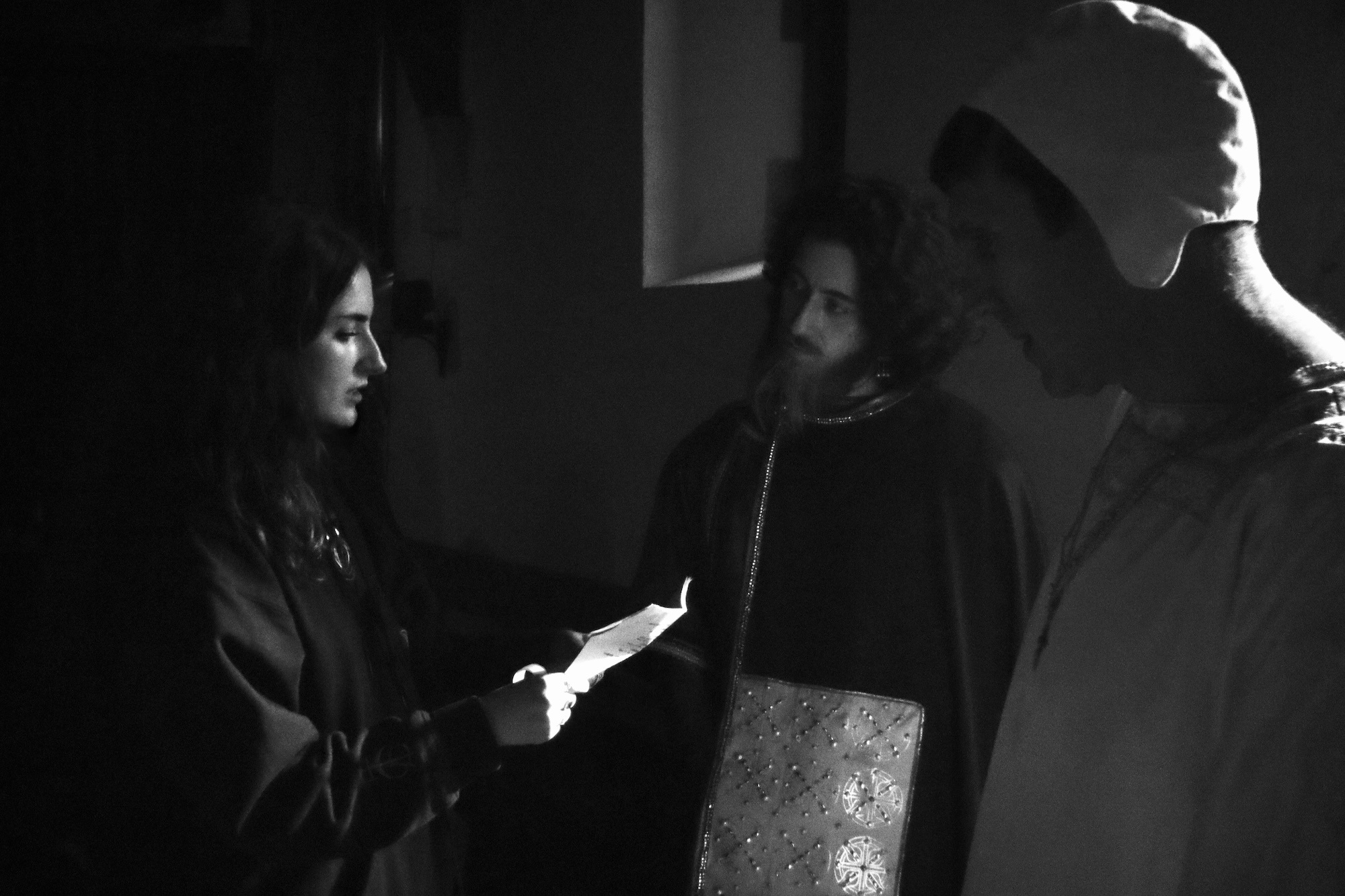 A woman in dark clothing holds a piece of paper while standing in front of two men, one with curly hair in a dark cloak and the other in a light-coloured garment. The scene is lit softly, creating shadows in a dimly lit environment.