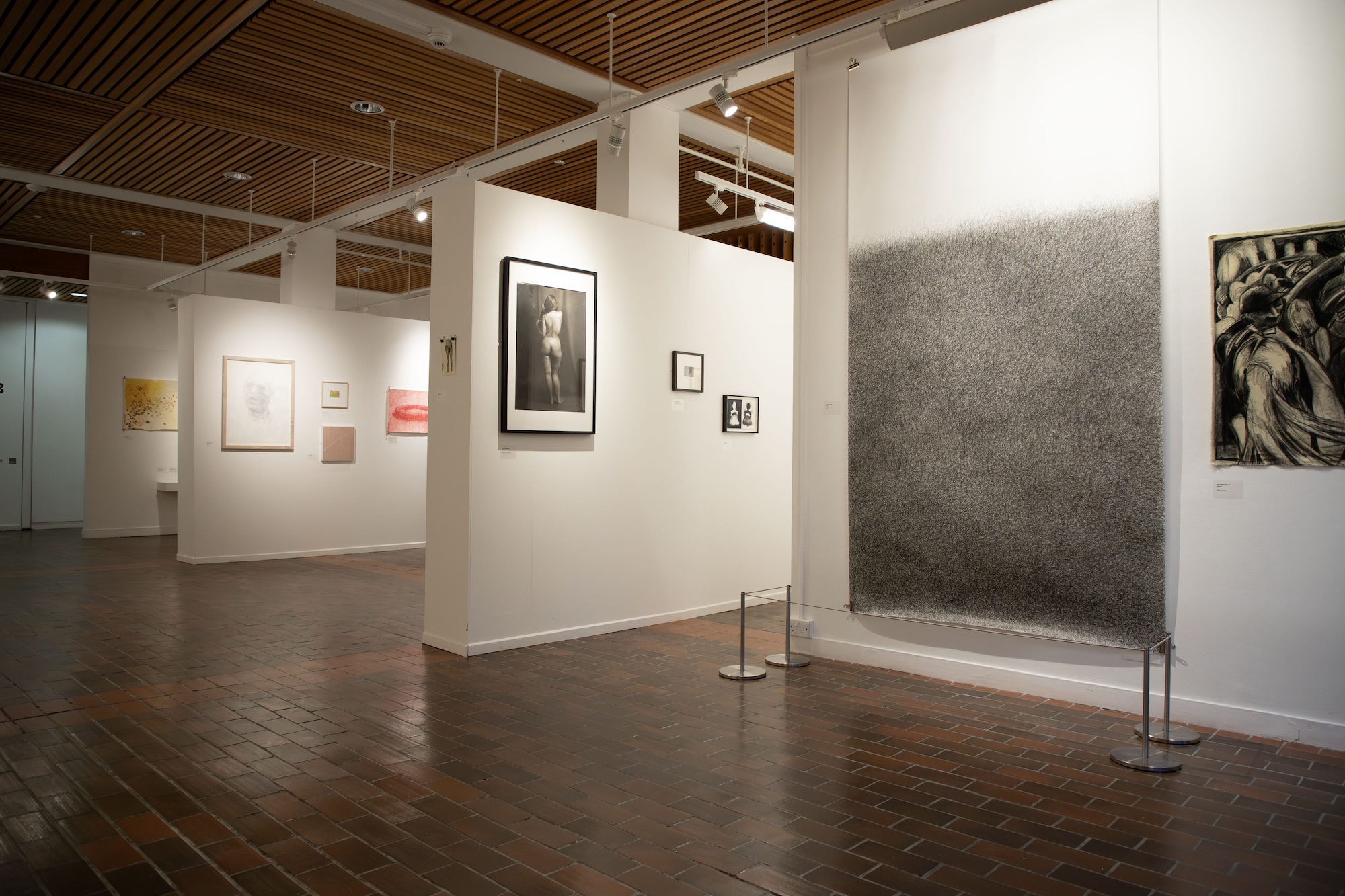 A contemporary art gallery featuring several framed artworks on white walls. To the right, a large dark charcoal drawing is displayed. The floor is tiled, and the space is well-lit with wooden ceiling beams.