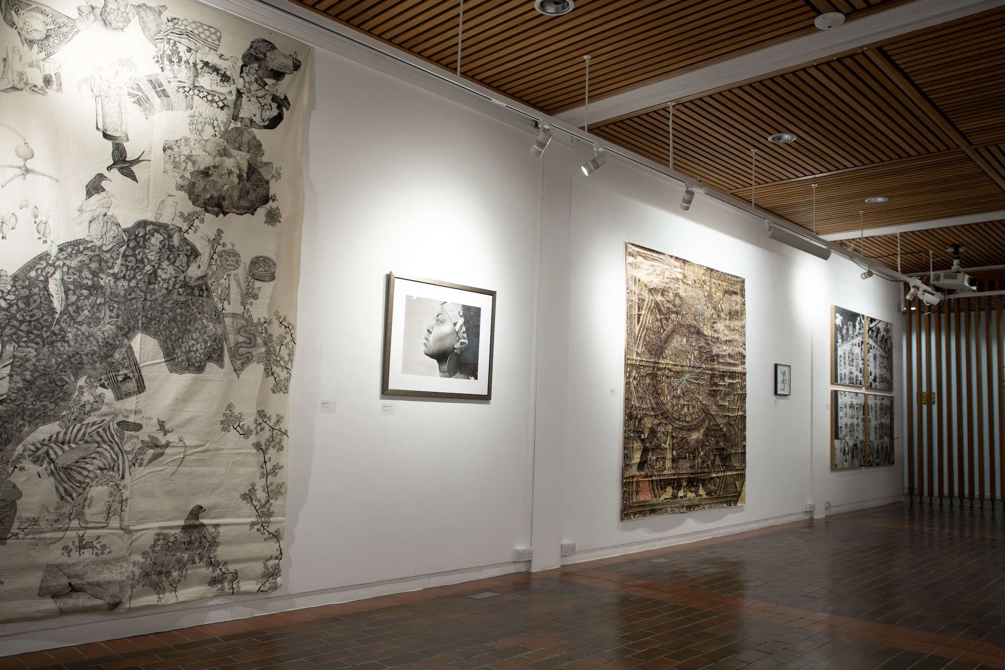 An art gallery showcasing four large artworks on white walls. On the left, a black and white fabric piece displays intricate patterns and motifs. In the centre, a framed monochrome portrait is displayed. The right side features two textured pieces of art.