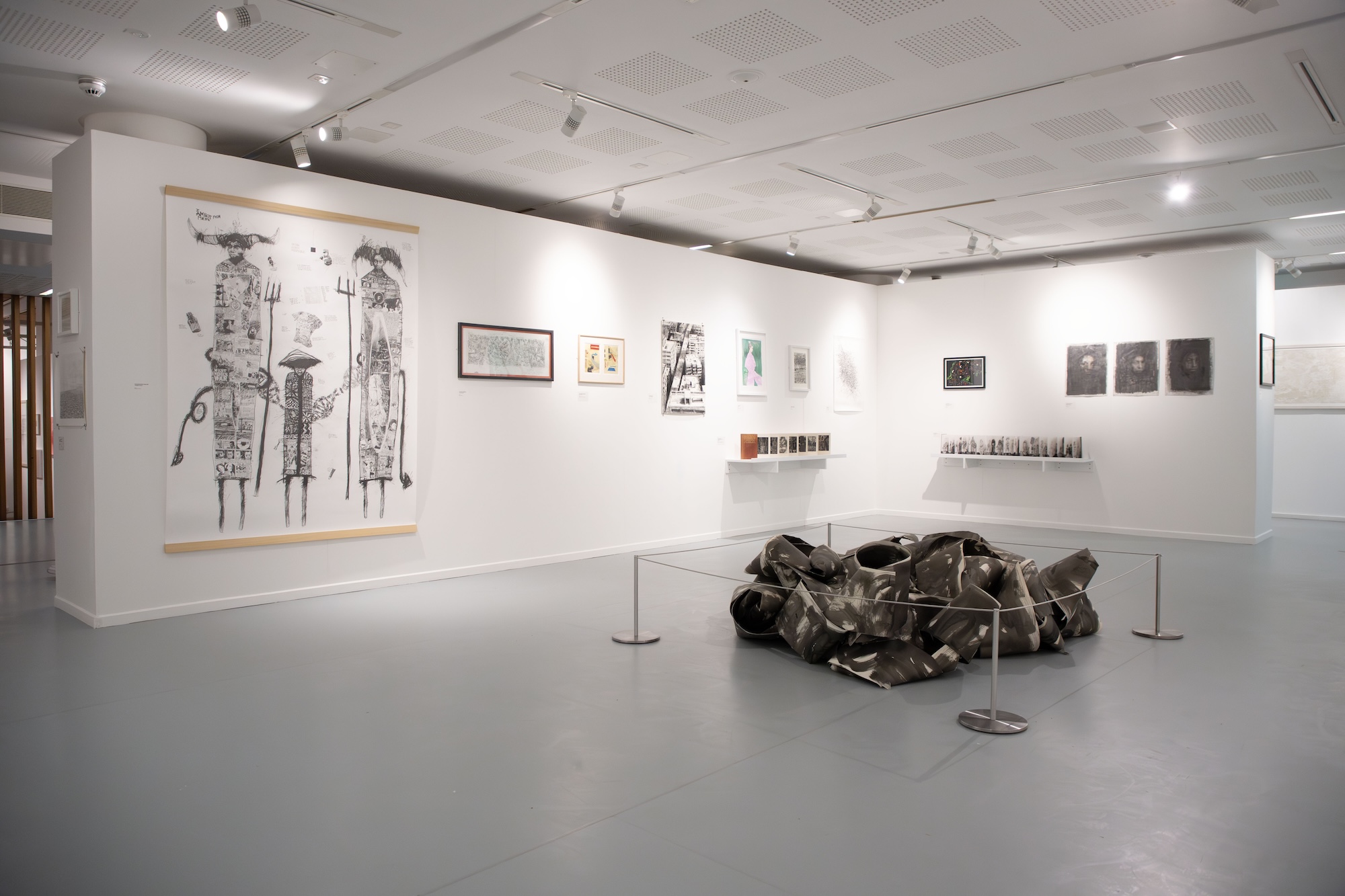 An art gallery interior featuring a large wall with various framed artworks, including illustrations and prints. In the centre, a sculptural installation made of dark, crumpled material is displayed, surrounded by barriers to the public.