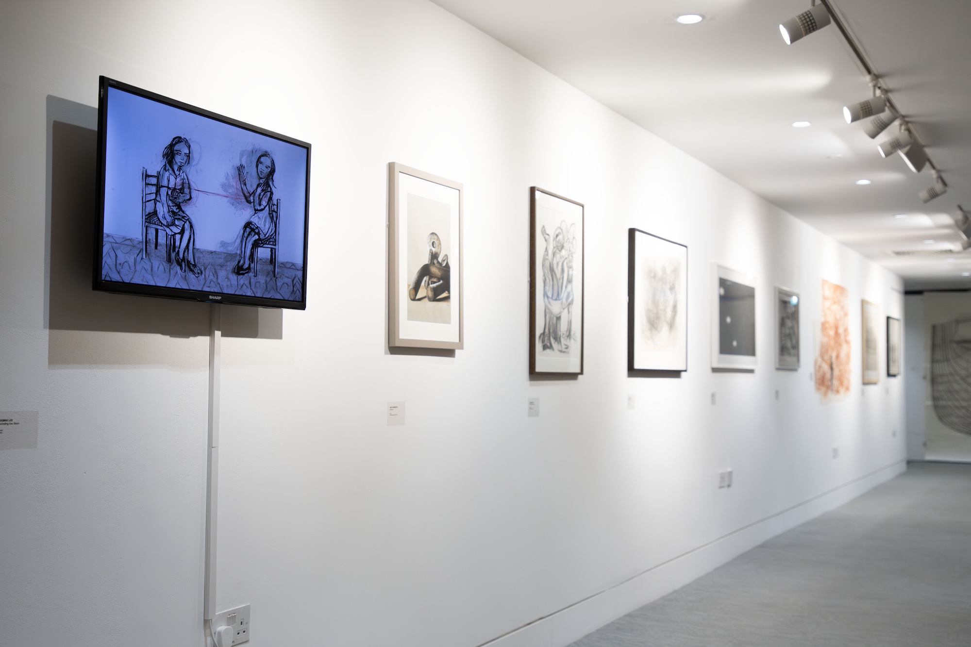 An exhibition space featuring a wall with various framed artworks. On the left, a television displays a sketch of two figures seated opposite each other. The artworks include drawings and illustrations, organised in a row along the white wall.