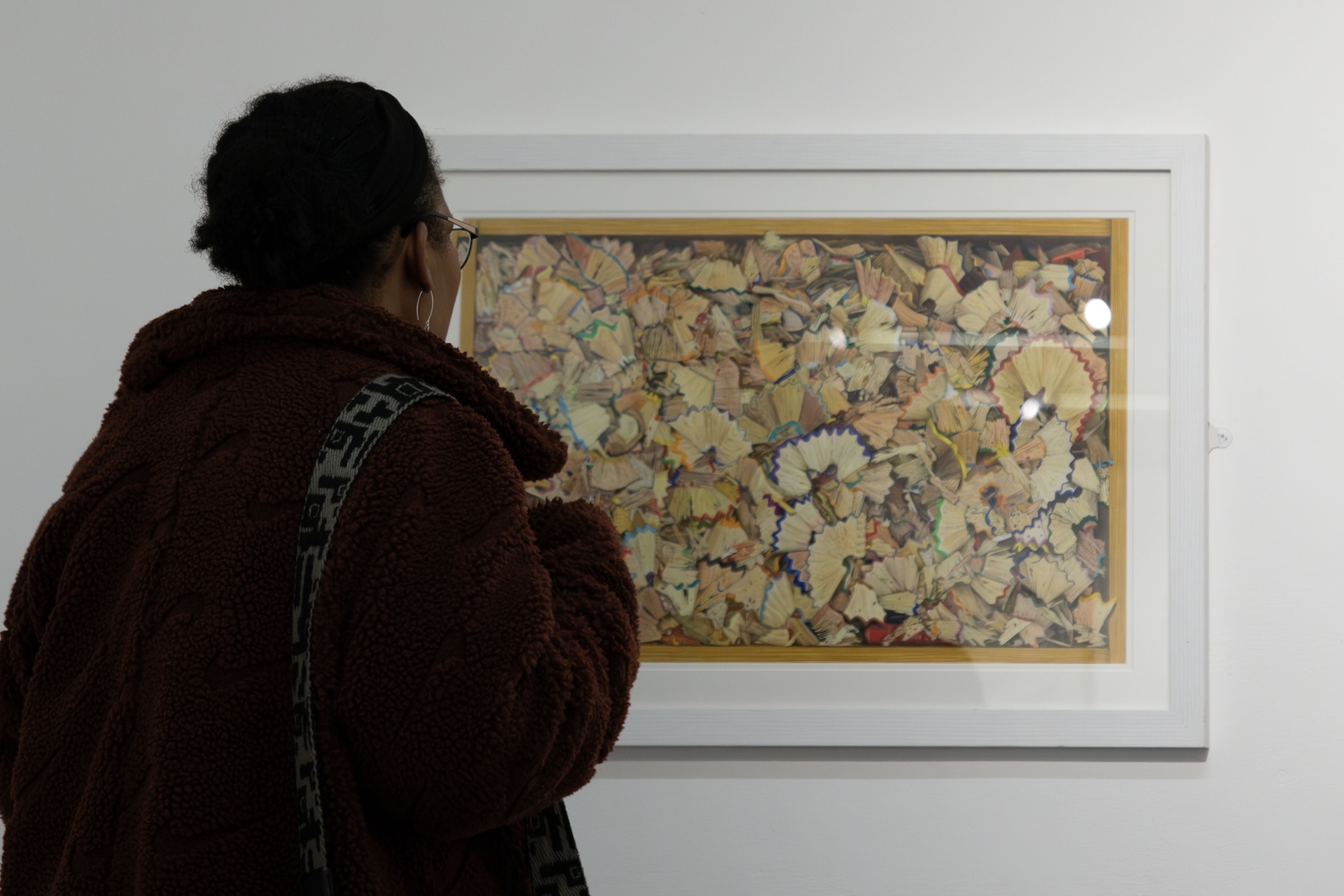 A person wearing a brown fluffy jacket stands in front of a framed artwork, which is a collage of various coloured leaves and shapes. The frame is white and enhances the vibrant colours of the artwork mounted on a plain wall.