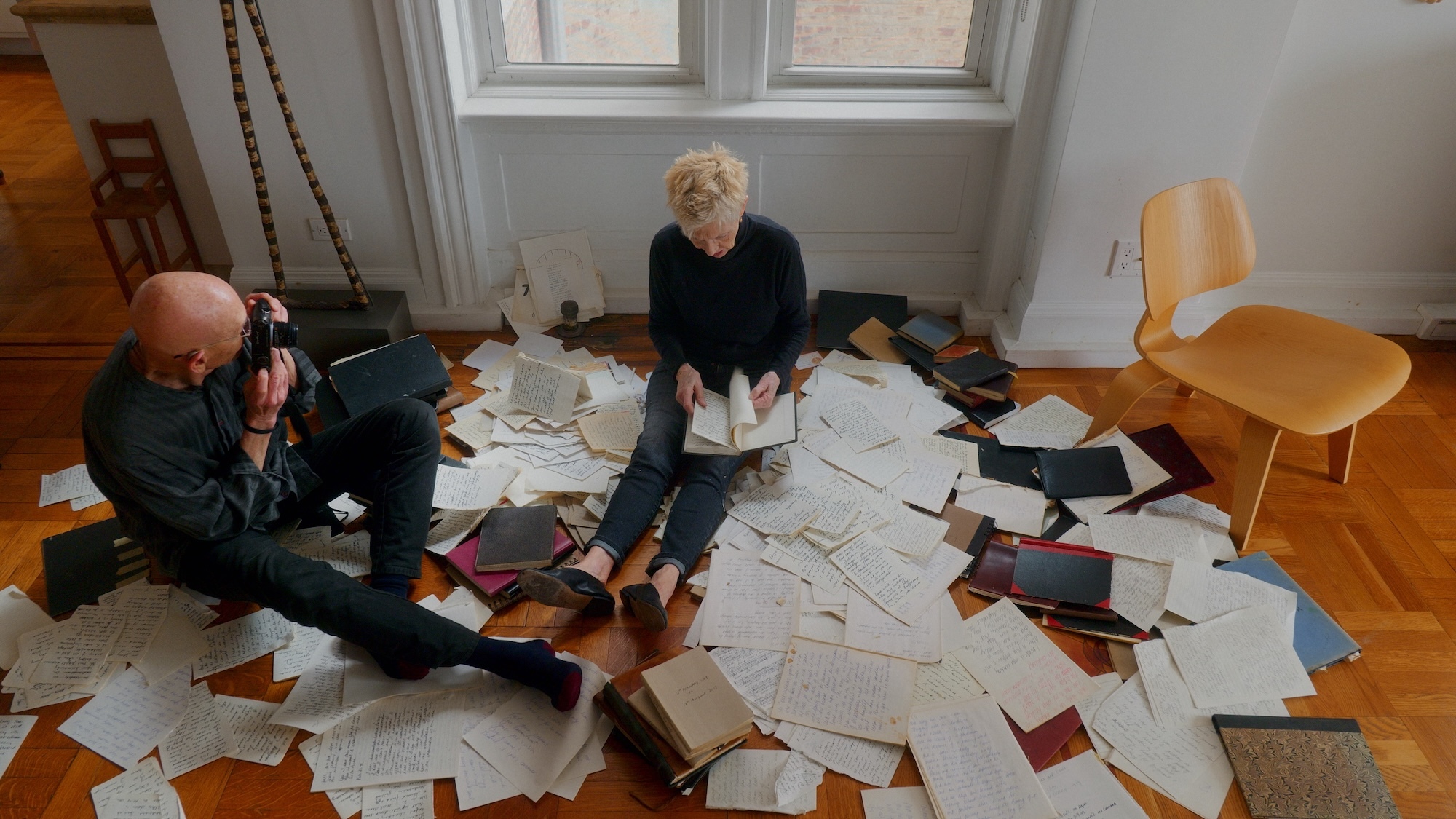 Still from "Two Strangers Trying Not to Kill Each Other". J Meyerowitz takes a photo of M Barrett as she sits on the floor surrounded by pages and journals.
