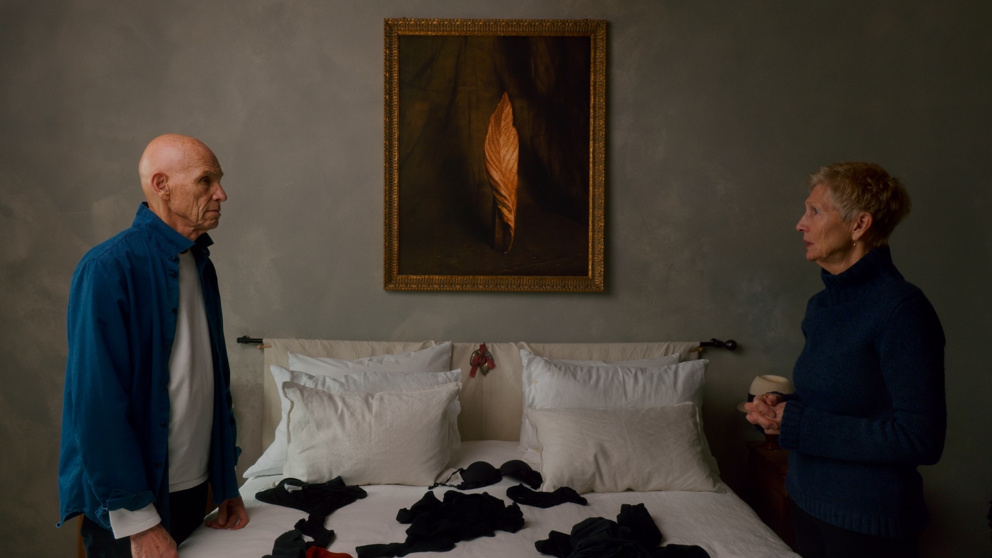 Still from "Two Strangers Trying Not to Kill Each Other". J Meyerowitz and M Barrett standing either side of a double bed facing each other, with black pieces of laundry strewn across the bed.