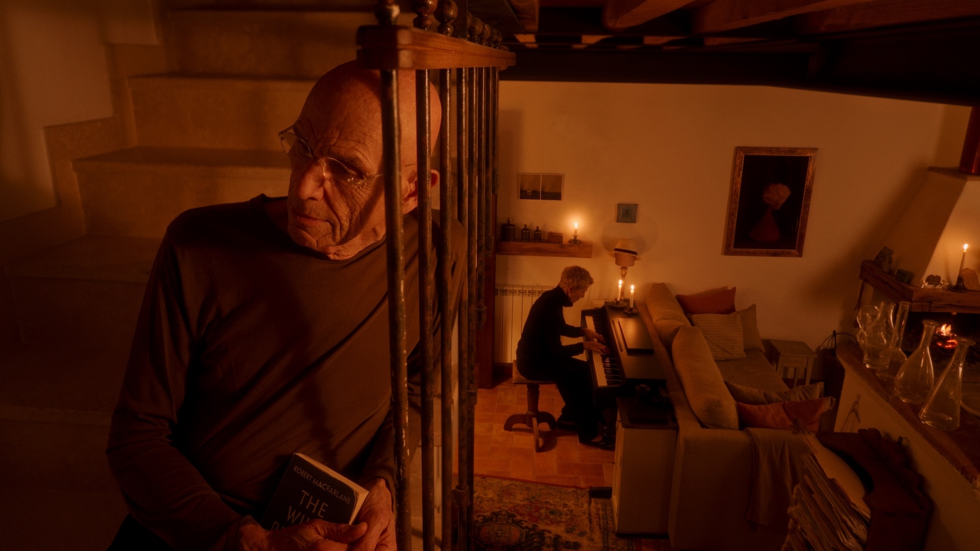 Still from "Two Strangers Trying Not to Kill Each Other". Close-up of J Meyerowitz sitting on some dimly lit stairs listening to M Barrett play the piano in the room below.