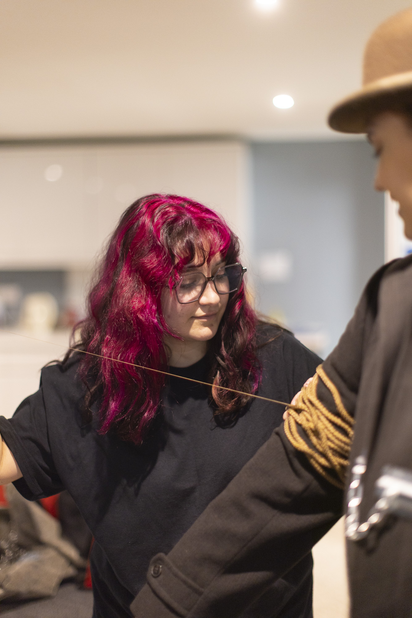 Woman with bright red hair and glasses wearing black top, partially obscured person in foreground, indoor setting with white walls.
