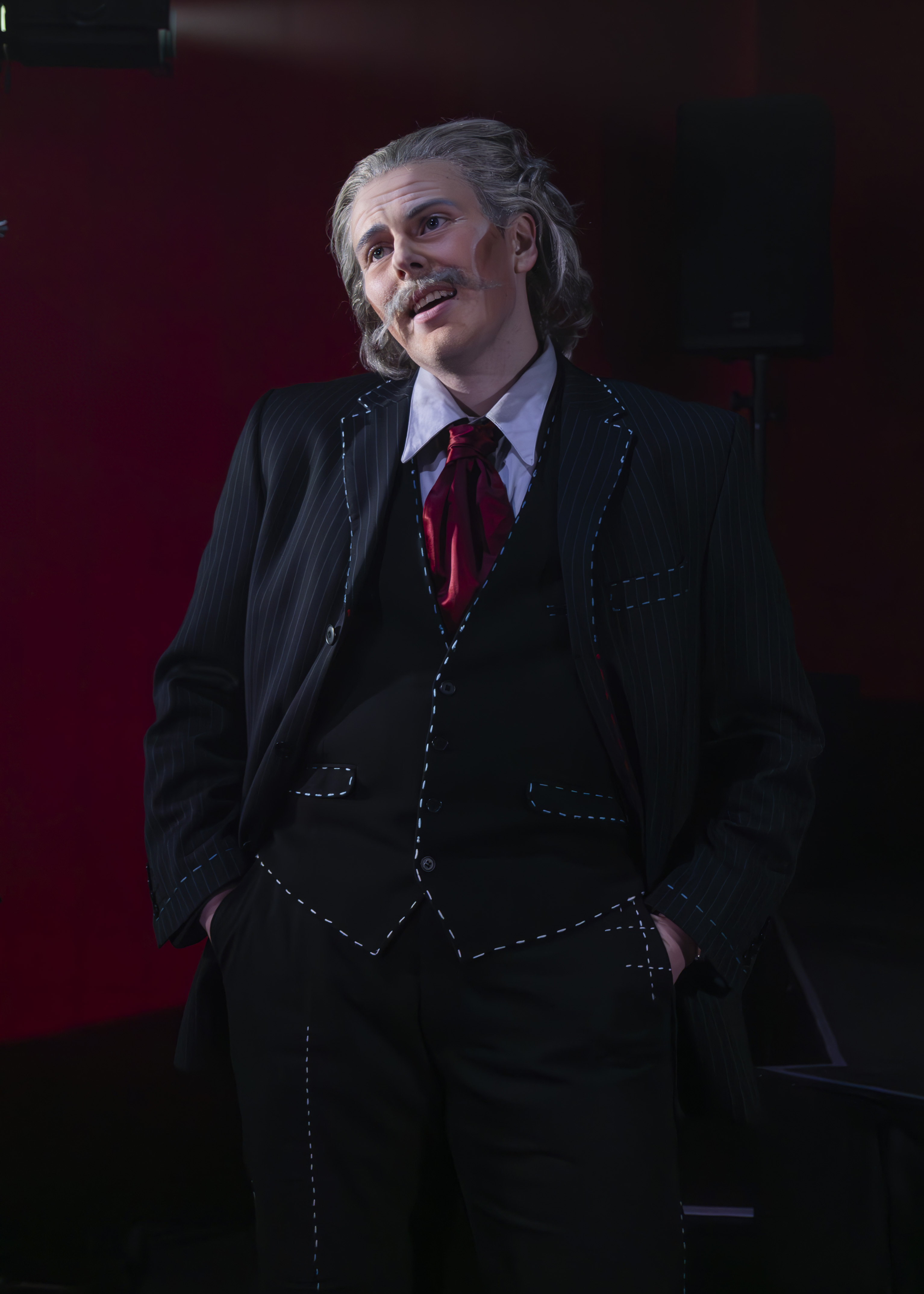 Actor in dark three-piece suit with red tie and waistcoat, grey hair and moustache, standing against burgundy background.