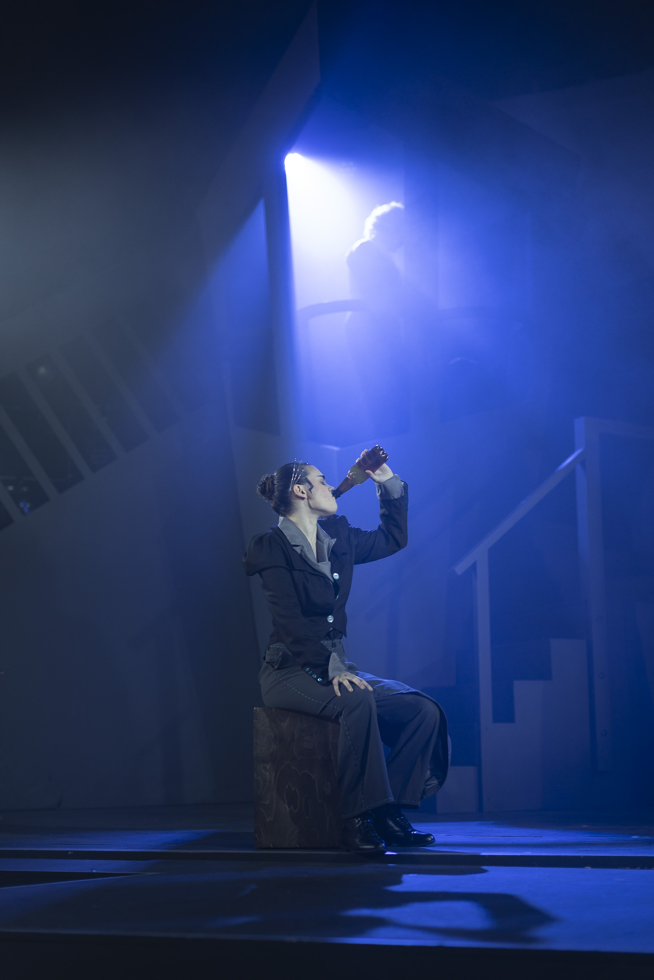 Performer in dark suit kneeling on stage, drinking from bottle under bright white spotlight with blue theatrical lighting.