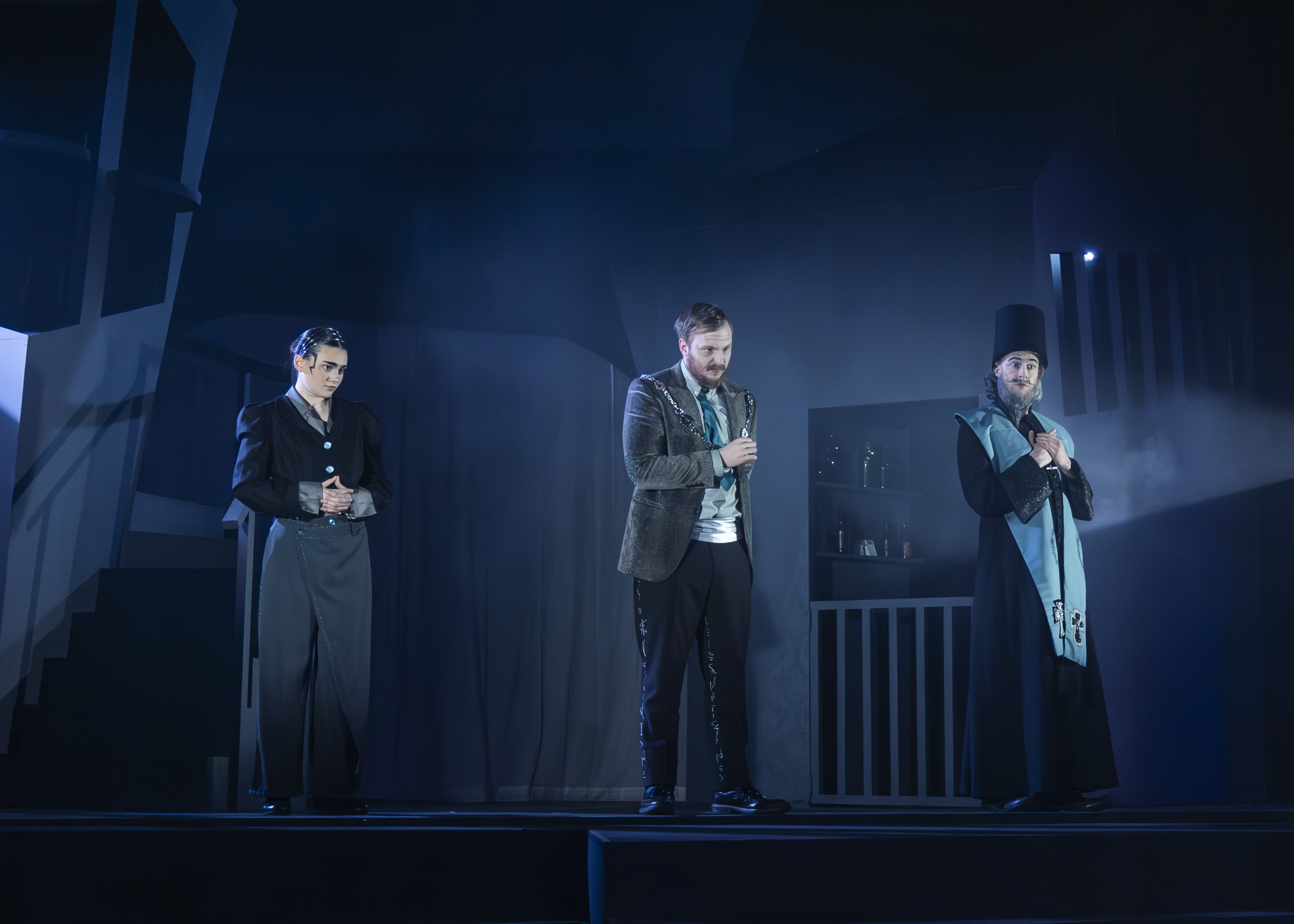 Three men in suits stand on a dimly lit stage with blue lighting and atmospheric fog.