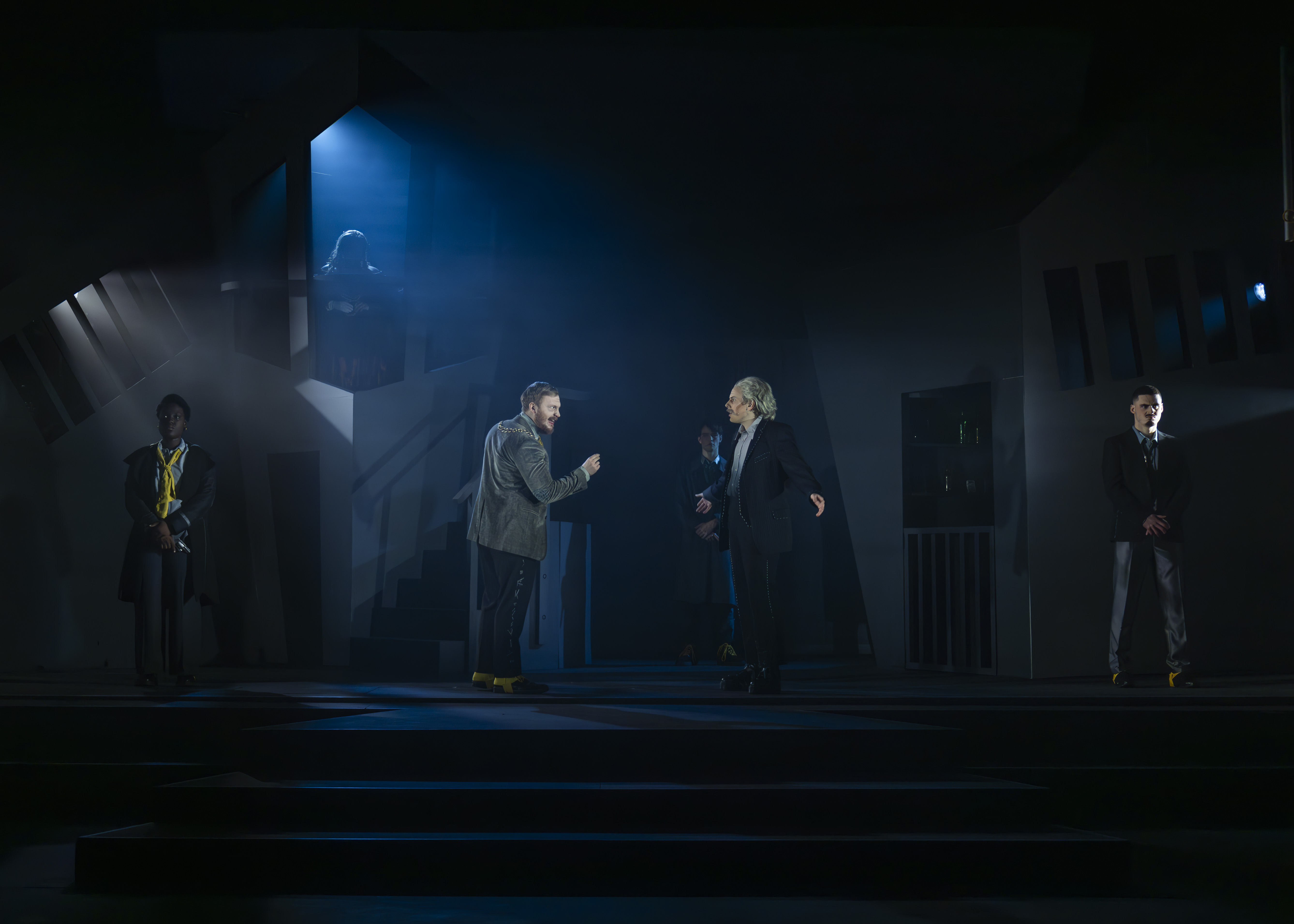 Theatre stage with dramatic blue lighting and smoke effects. Four actors in dark clothing positioned across the stage, with bright spotlights creating atmospheric beams through the haze.