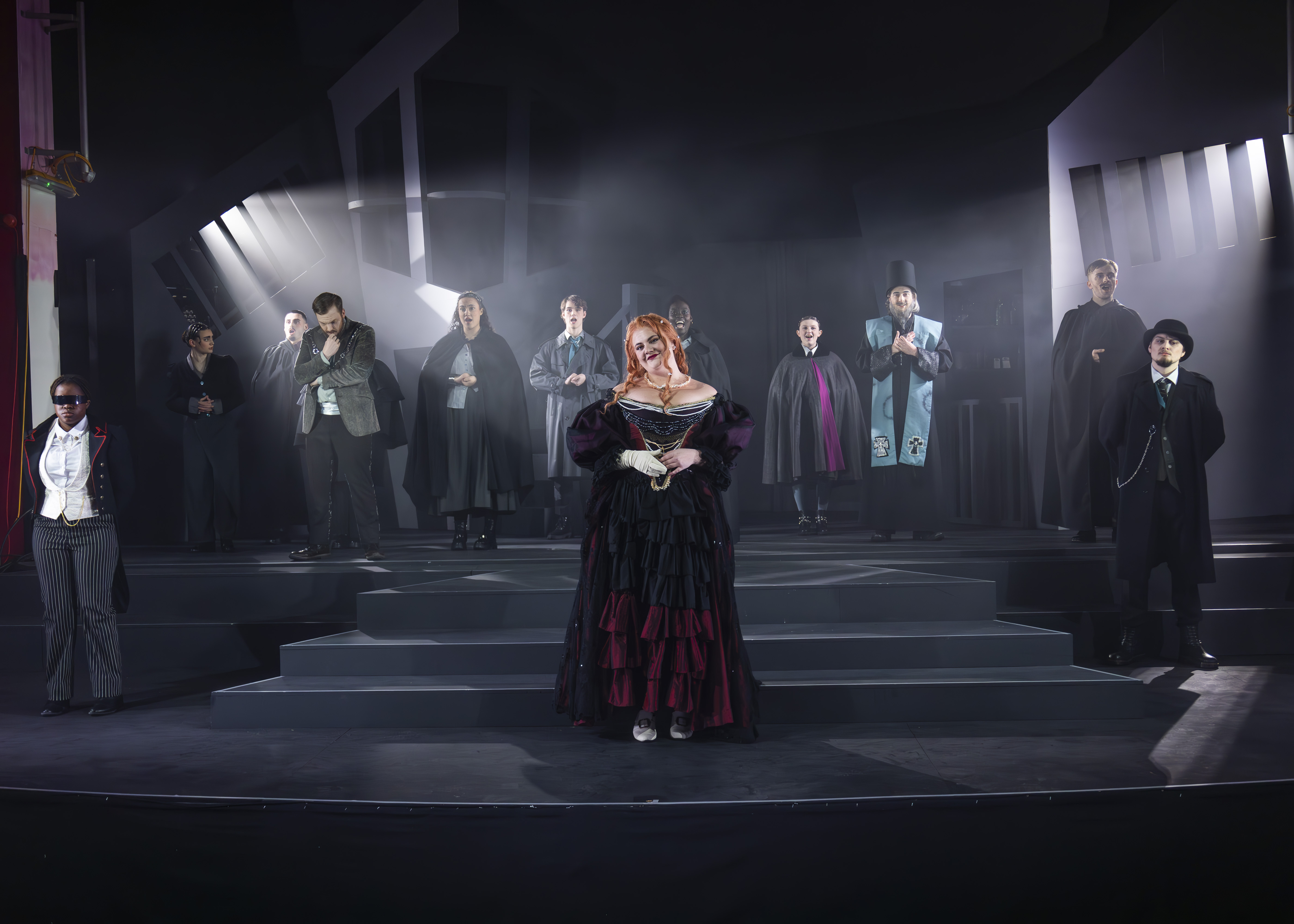 Theatre stage with performers in period costumes, woman in burgundy dress centre stage, dramatic lighting with white beams.