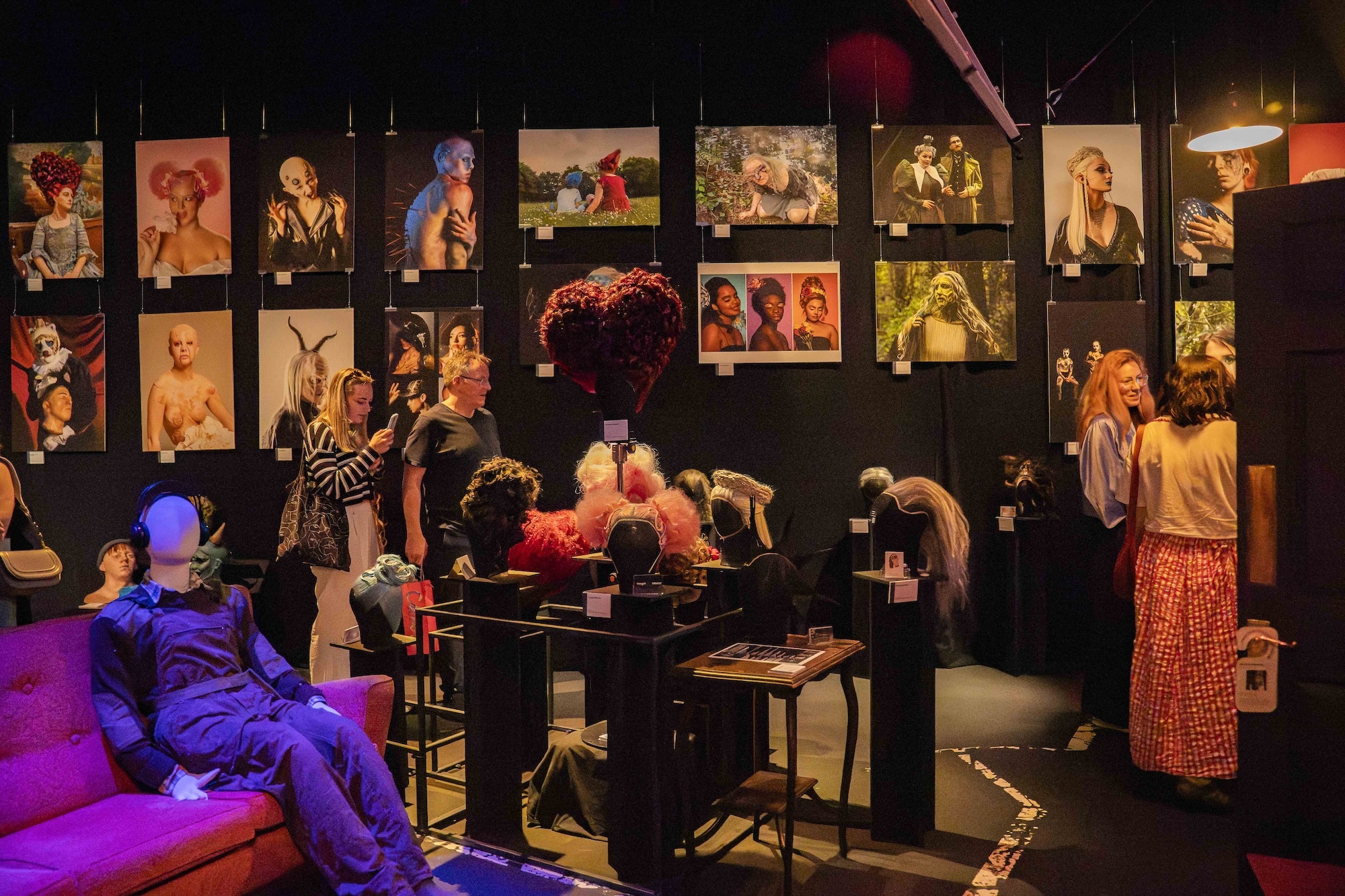 People exploring a black studio room filled with various portraits of different hair and make-up looks. Wigs are on display on a table in the centre of the room. A mannequin sits on a red sofa to the left-hand side.