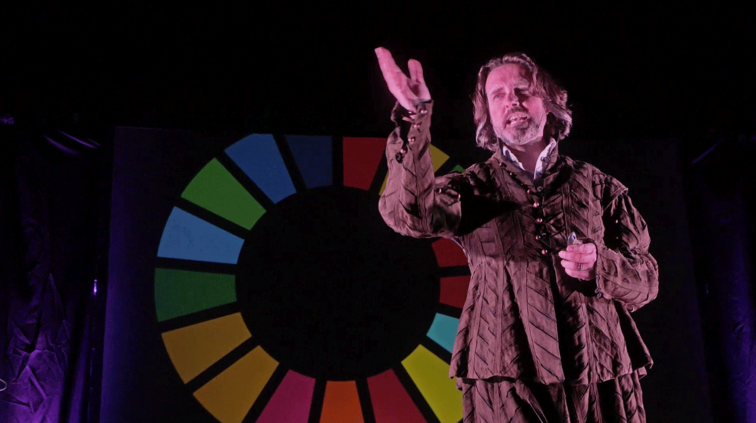 A man performing in costume on a stage with a circle of colours projected in the background.