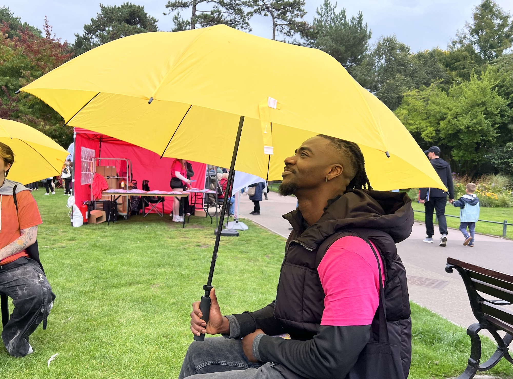 A man in a pink T-shirt and black vest sits on a bench holding a large yellow umbrella in a park, with a pink tent and other people in the background.