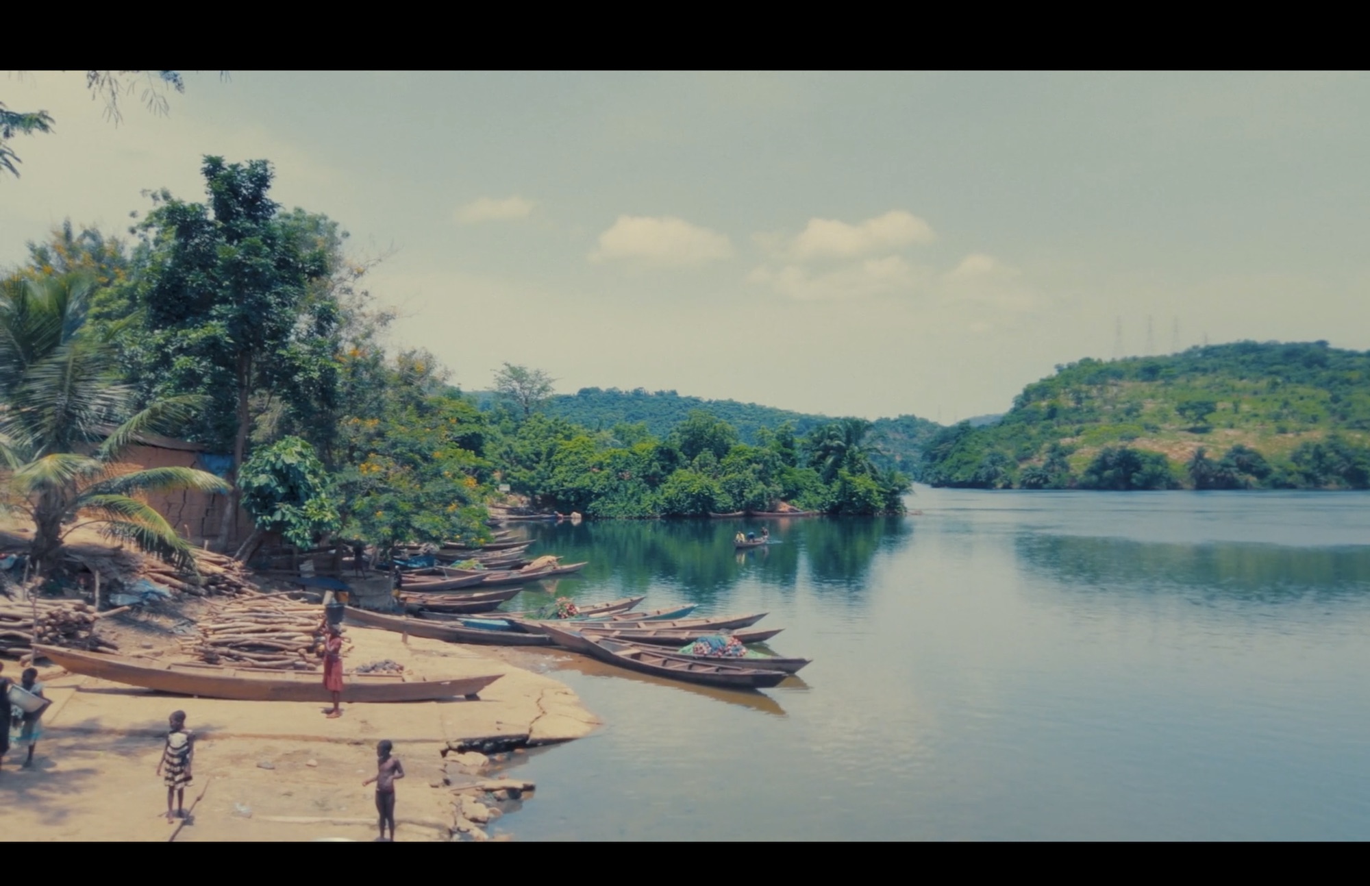 A scenic riverbank with several wooden boats moored along the shore. In the foreground, children play on a sandy area, while lush greenery and hills rise in the background under a clear blue sky.