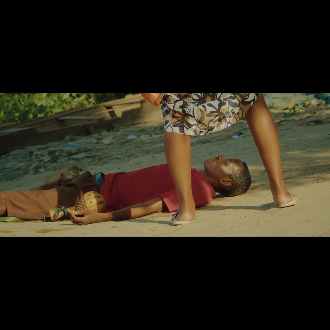 Woman in patterned shorts standing over man lying on beach sand in red shirt, with water and greenery in background.