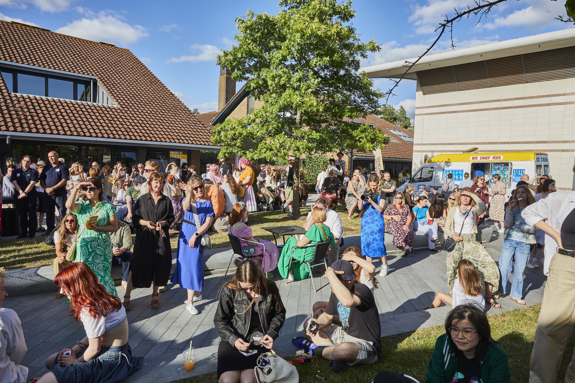 Large crowd gathered in the courtyard between the North Building and Library on Arts University Bournemouth campus, under blue sky with clouds.