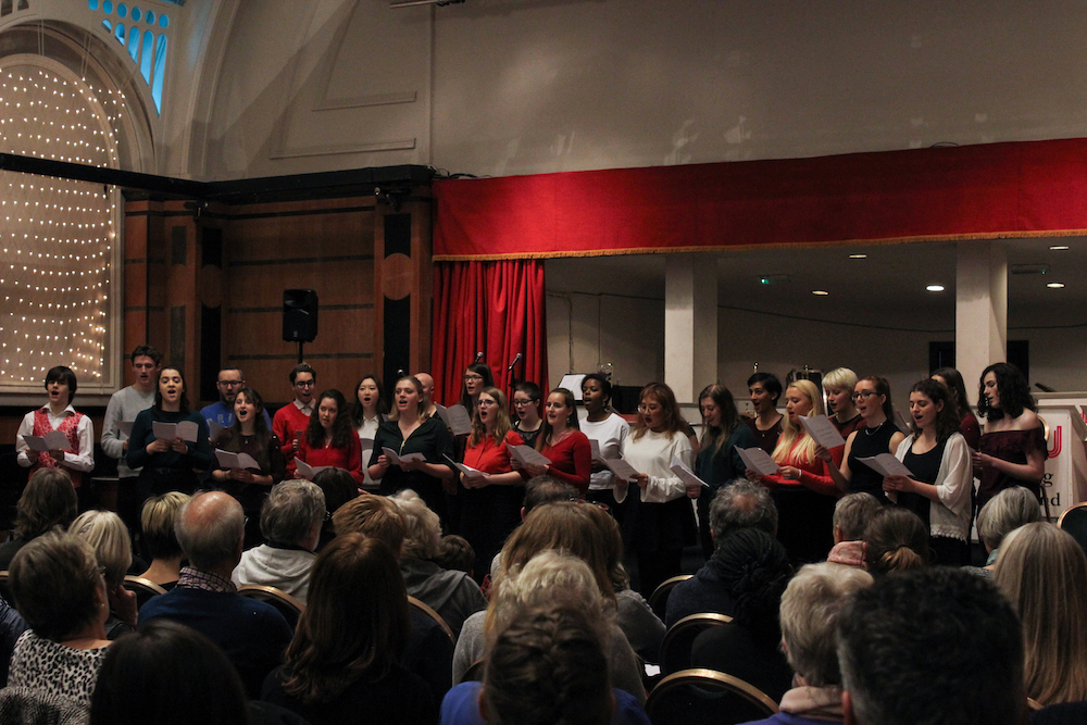 A choir stands in two rows and sings before an audience in a theatre.