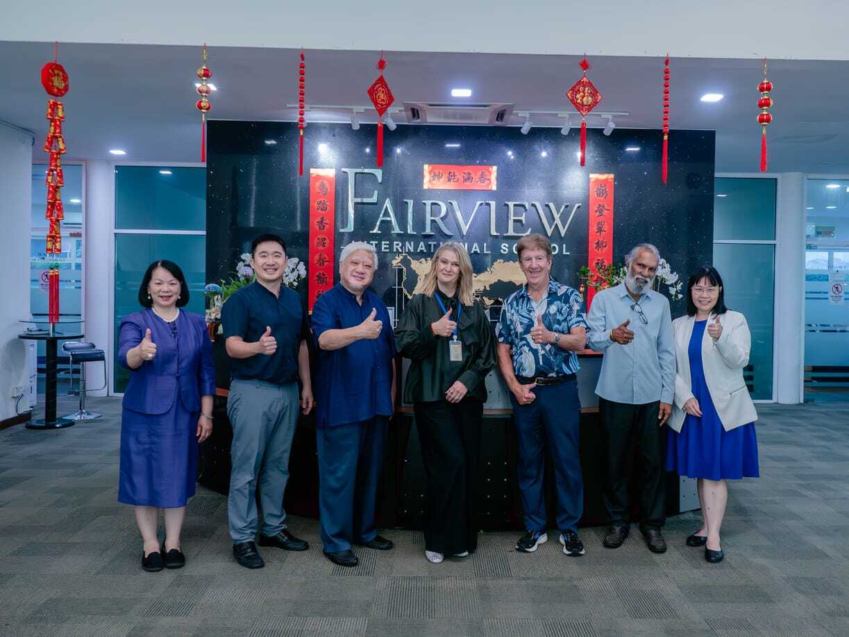 Group of eight people posing in office lobby with Fairview International School sign and red Chinese New Year decorations hanging.