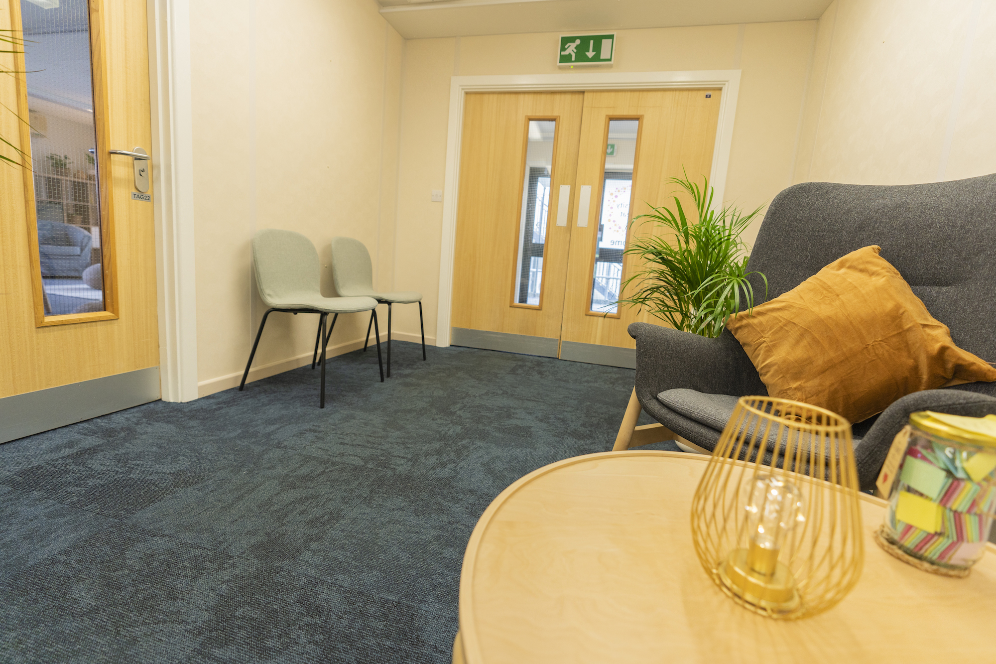 A waiting room with a coffee table and armchair on the right. Another pair of chairs stand in a corner between two sets of doors.