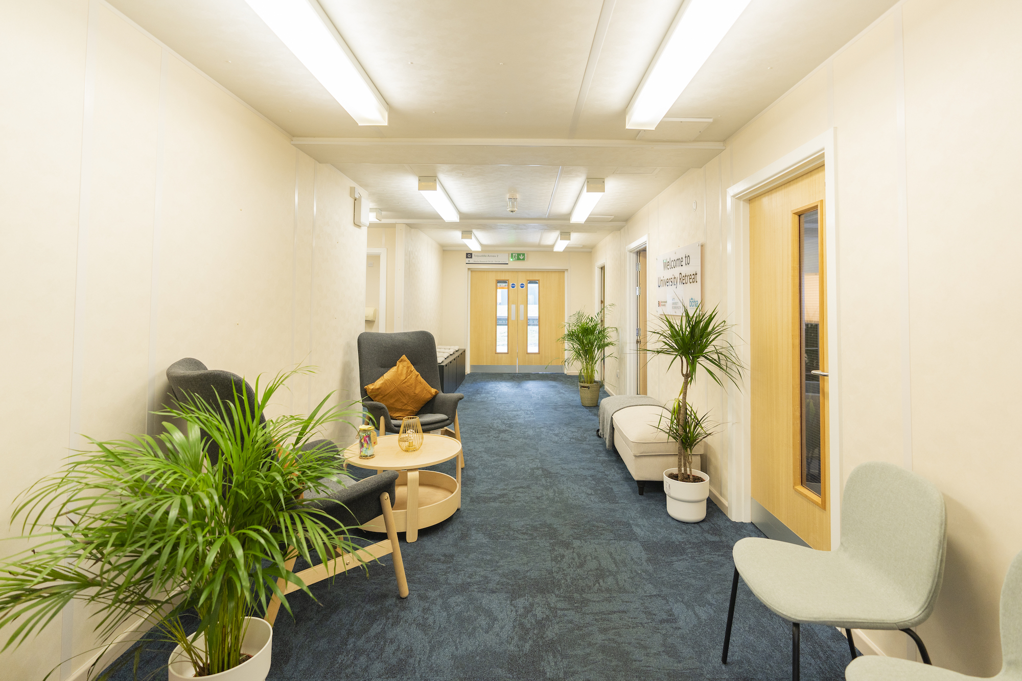 Waiting room with chairs and a coffee table on the left, more chairs and three doors on the right. A pair of double doors are down the far end of the room.