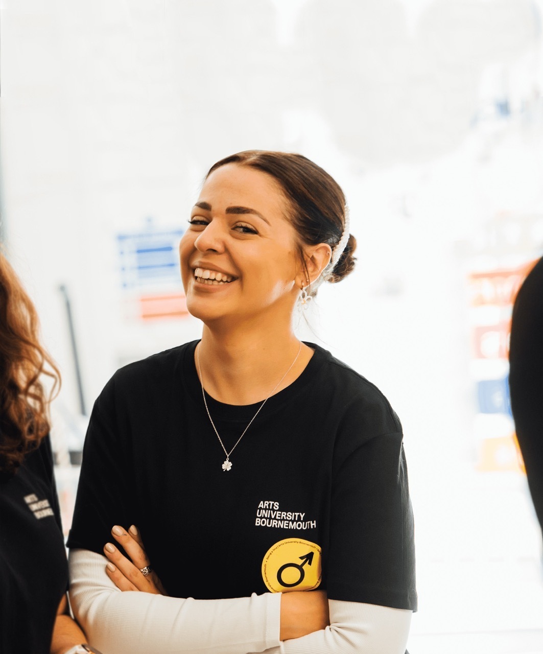 Woman with hair in bun wearing black Arts University Bournemouth t-shirt with yellow circular logo, smiling in bright indoor space.