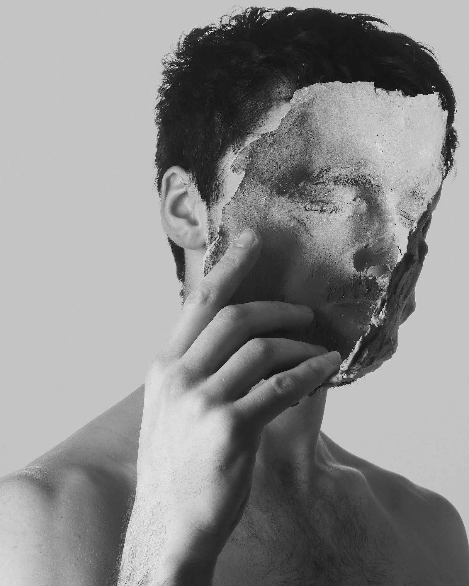 Black and white head and shoulders shot of a topless man holding a torn photograph in front of their face. The photograph shows another face with its eyes closed.