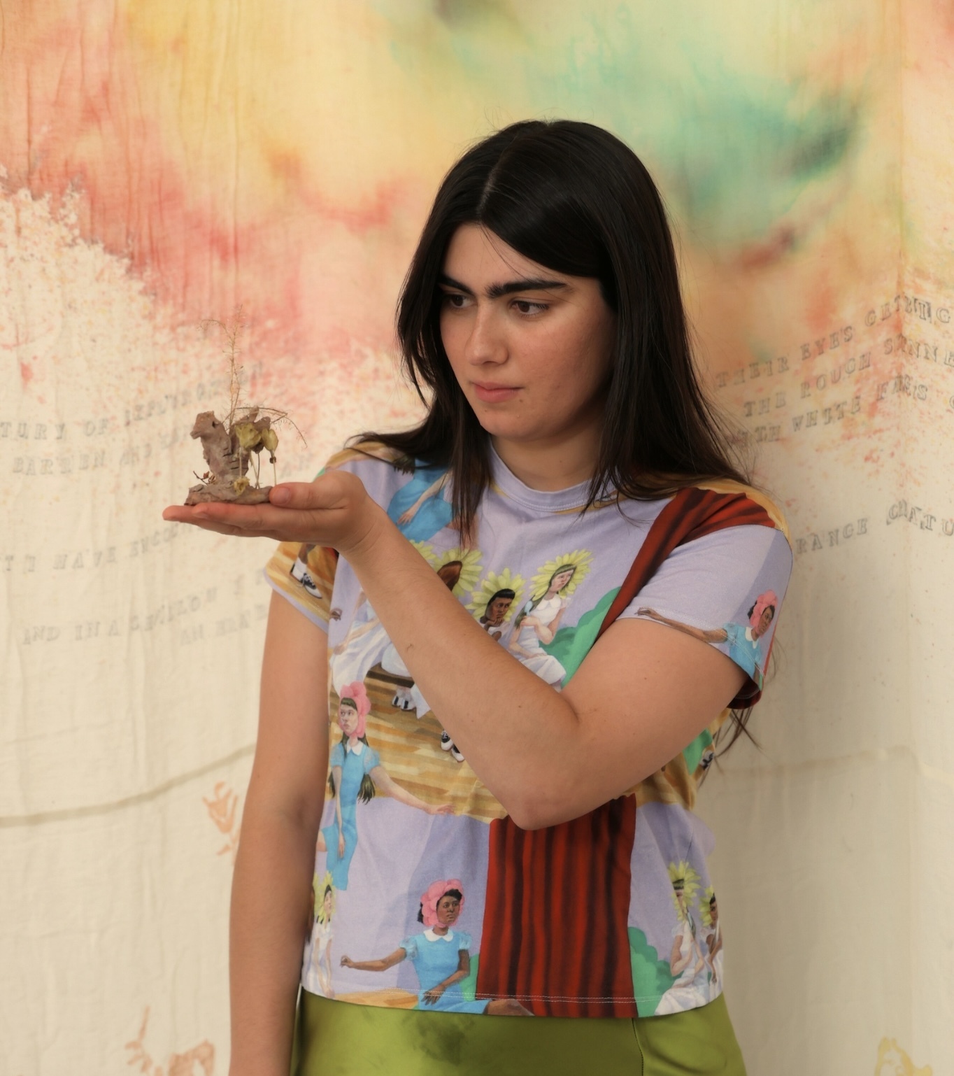 Woman with dark hair holding small object, wearing colourful patterned top and green trousers against rainbow watercolour background.