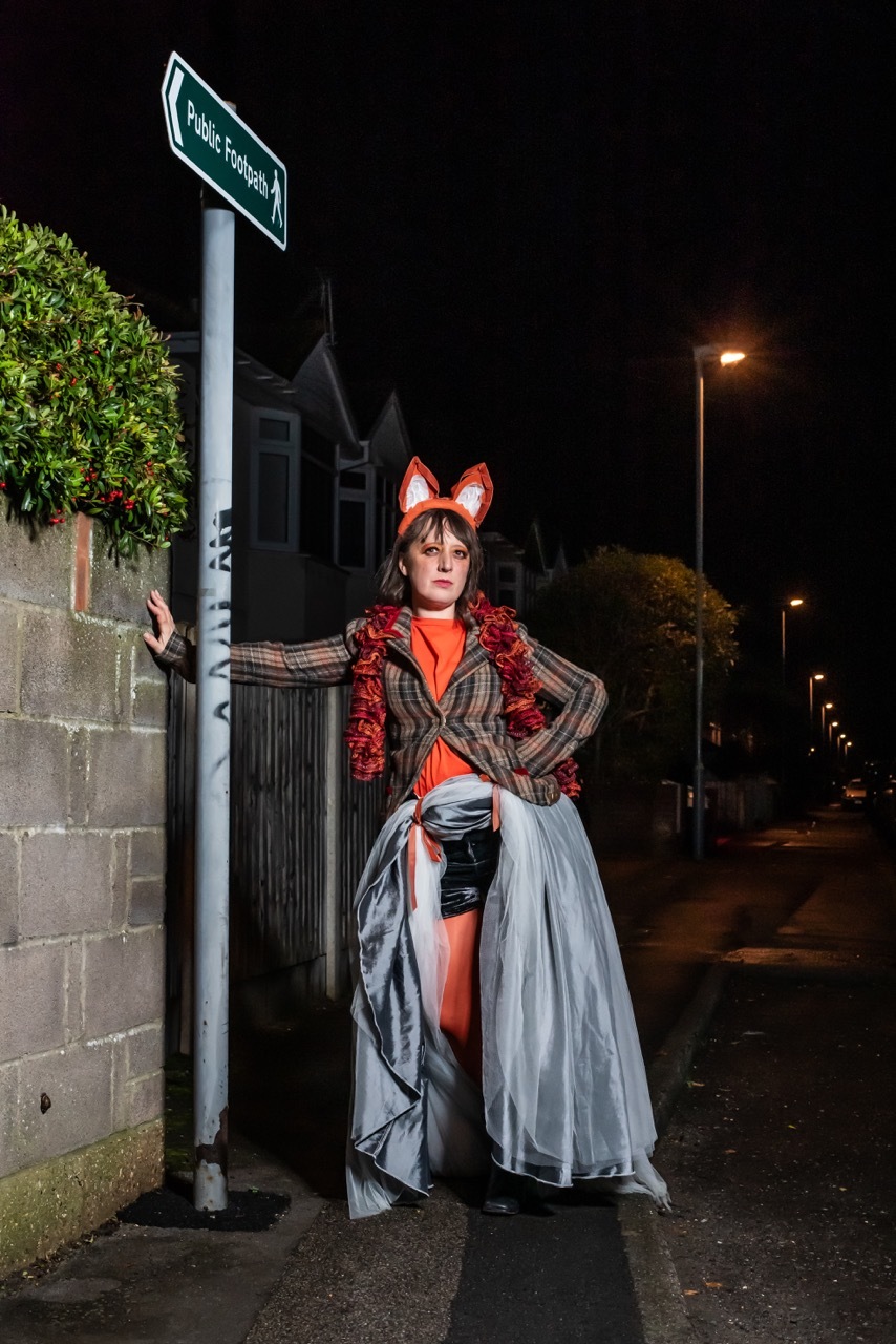 A woman standing on a street at night, dressed in fox ears and orange, extravagant clothing.
