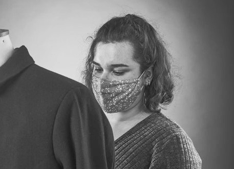Black and white image of person with shoulder-length dark hair wearing patterned face mask, holding dark clothing item.