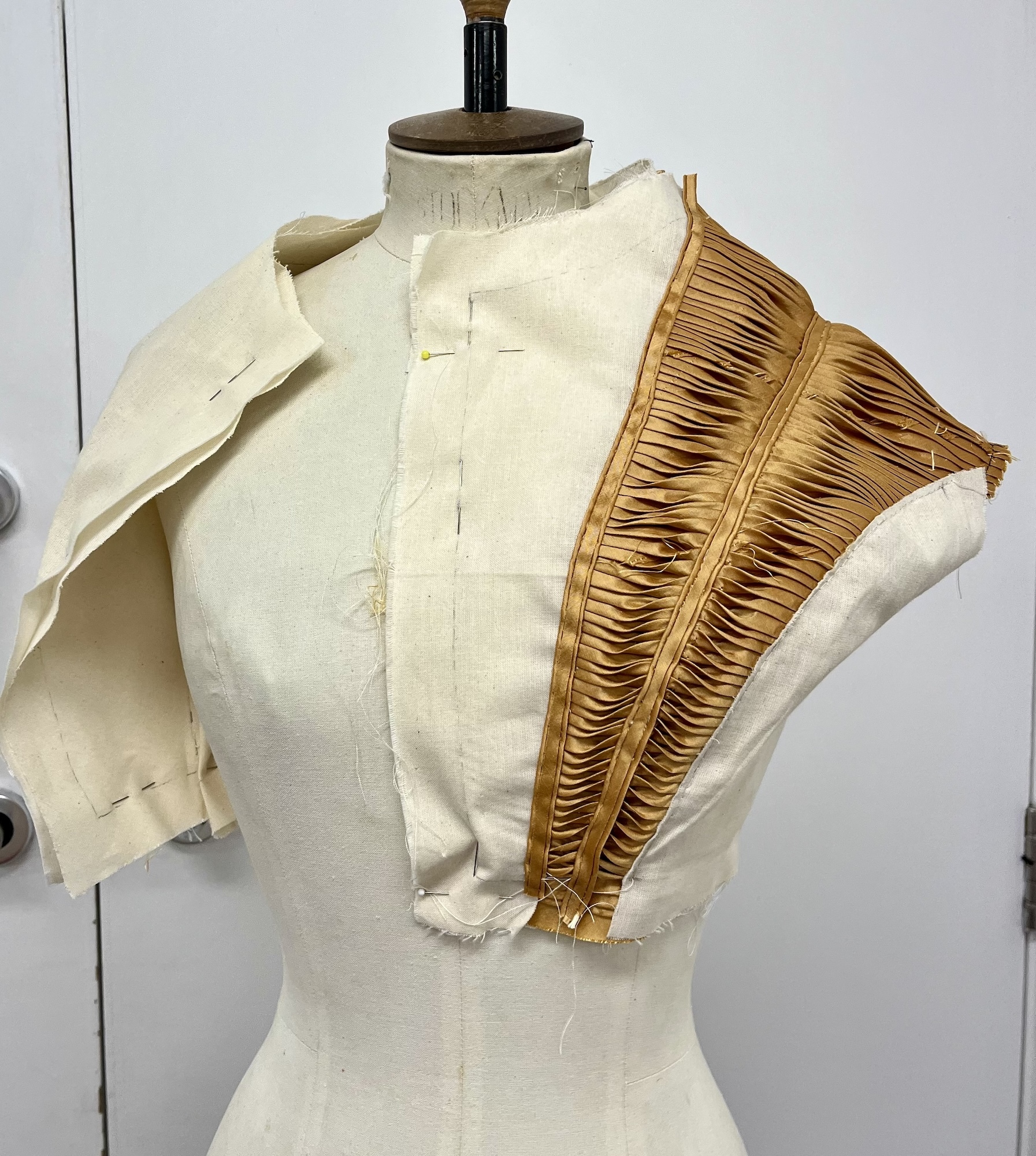 A mannequin with a piece of gold-coloured material pinned in place across the left shoulder and part of the chest.