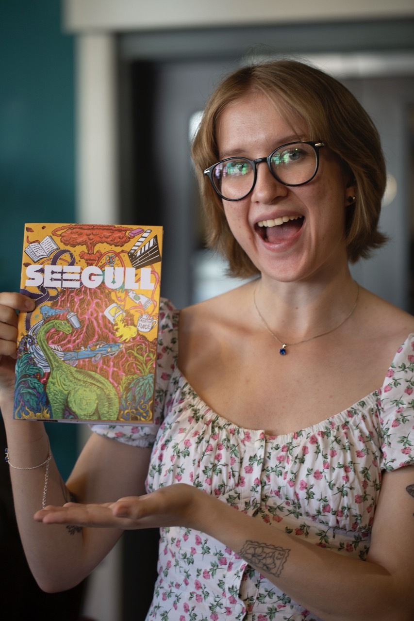 Maria Bishop with acopy of Seegull magazine, title in white with a vibrant illustration of a volcano, dinosaur, book and other creative items on a yellow background.