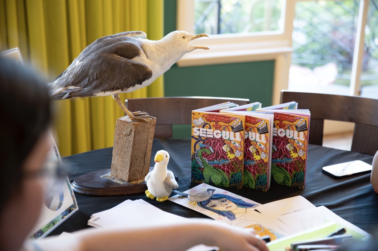SEEGULL launch event with a stuffed seagull and plush toy with copies, title in white with a vibrant illustration of a volcano, dinosaur, book and other creative items on a yellow background.