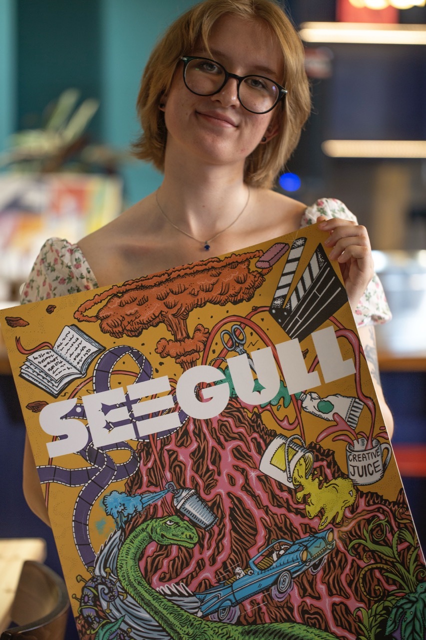 Maria Bishop with a large poster of the Seegull magazine front cover, title in white with a vibrant illustration of a volcano, dinosaur, book and other creative items on a yellow background.