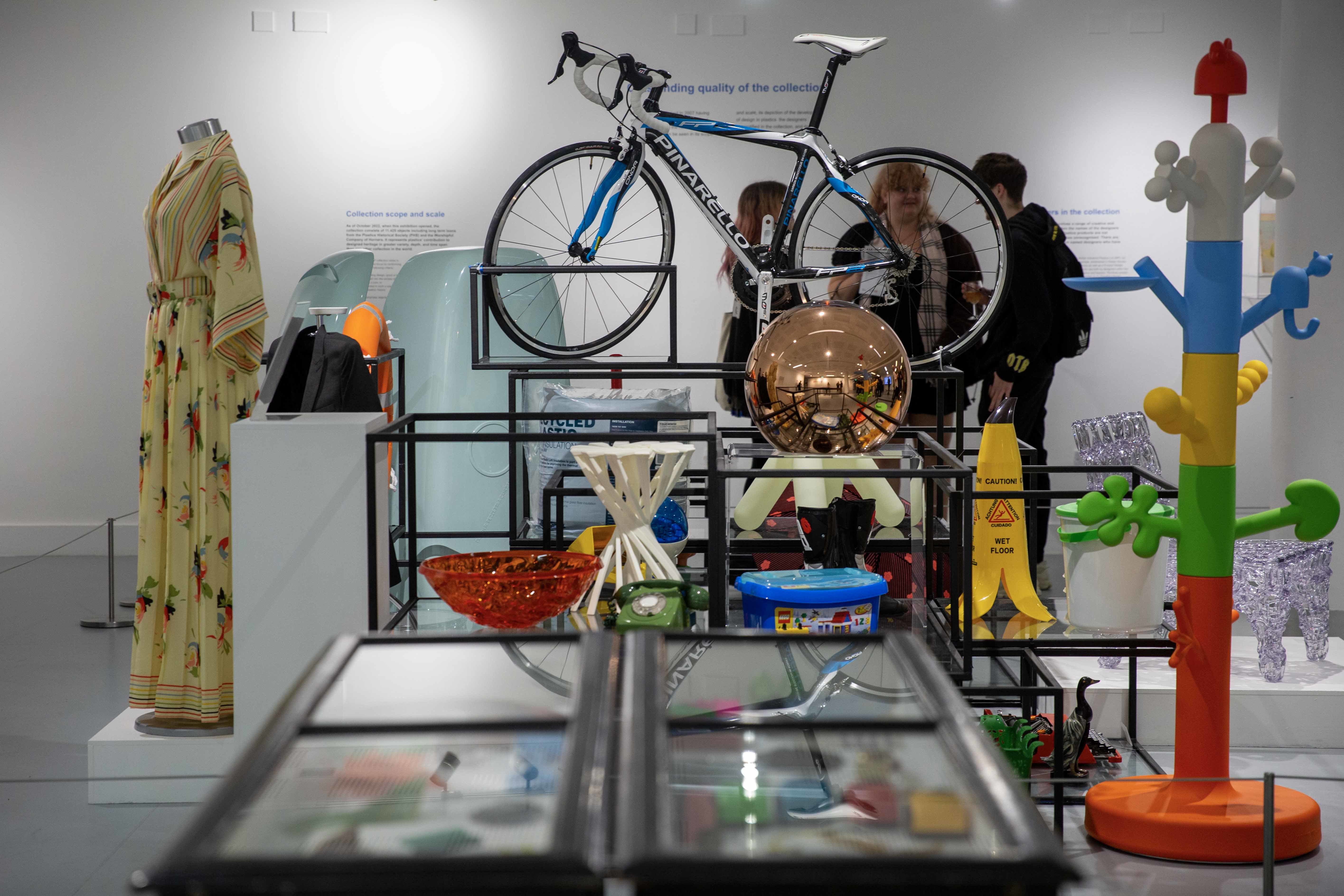An assortment of exhibits on display in a gallery space, including a bicycle, a colourful plastic hat rack, a box of Lego and a fruit bowl.