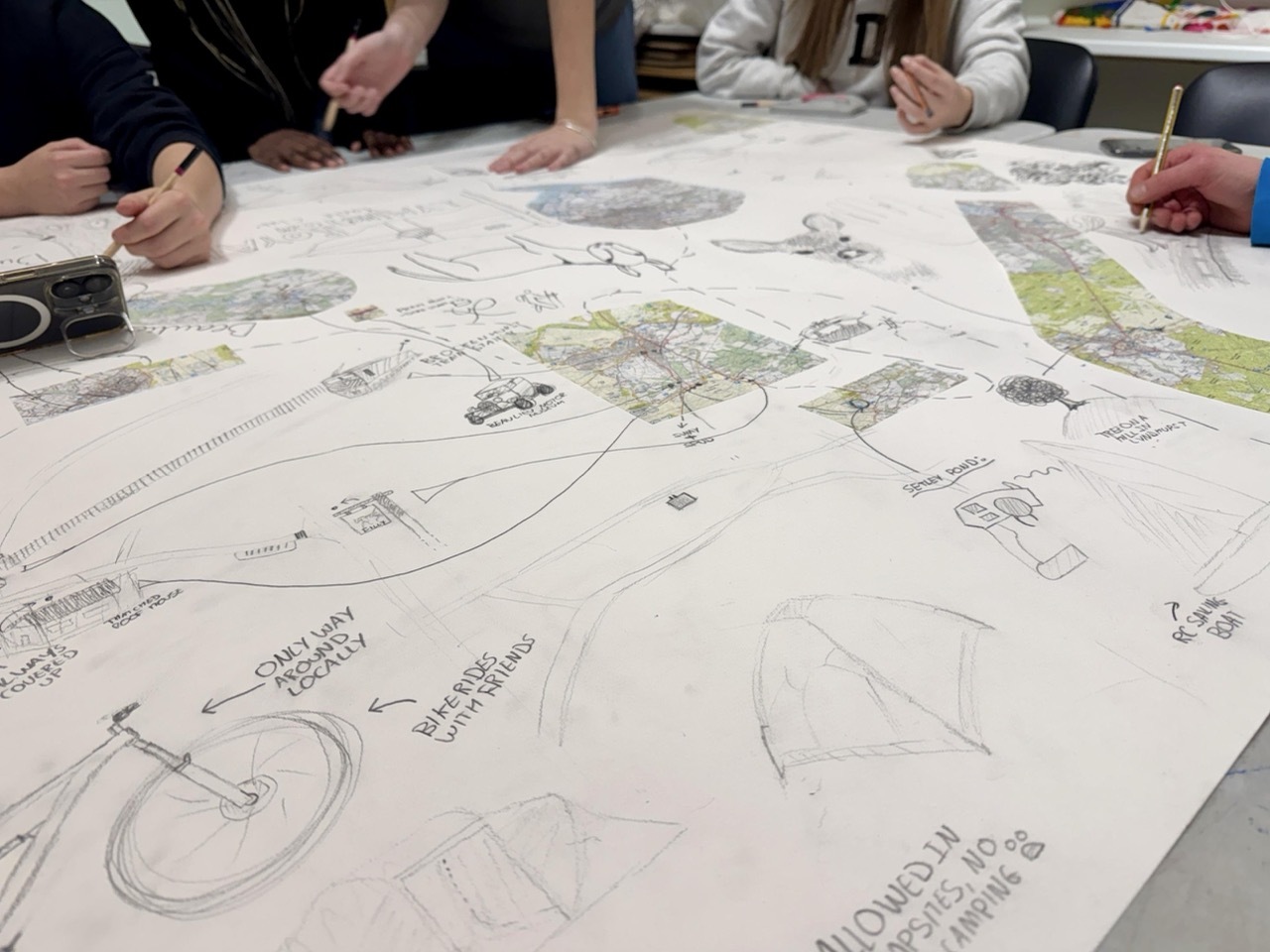 People gathered around large architectural plans and drawings spread across a table, hands visible pointing at and discussing the designs