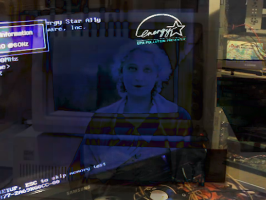 A CRT monitor displaying a blue-tinted woman’s portrait, with reflected interface text and a logotype in the upper right corner.