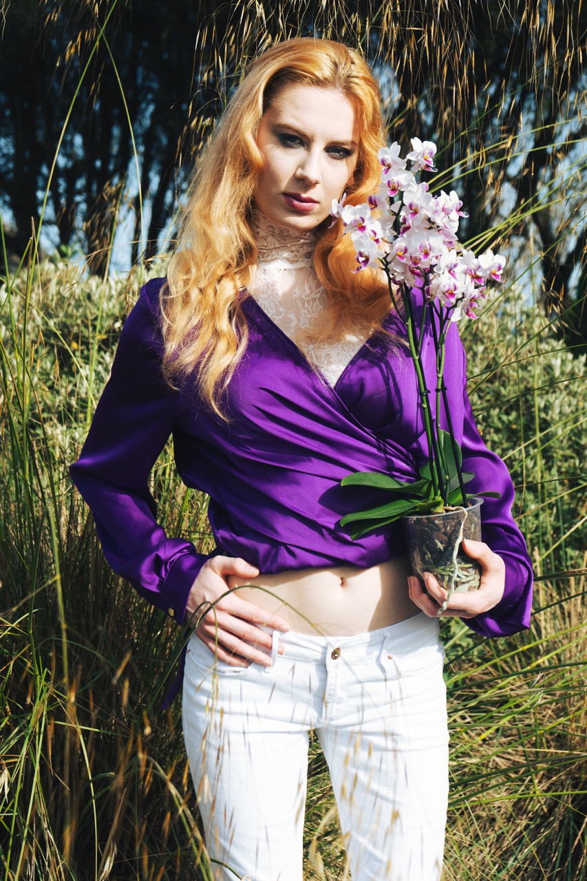 A woman with long blonde hair wears a deep purple blouse and white trousers, holding a pot of pink orchids. She stands against a backdrop of tall grass and trees, with sunlight highlighting the scene.