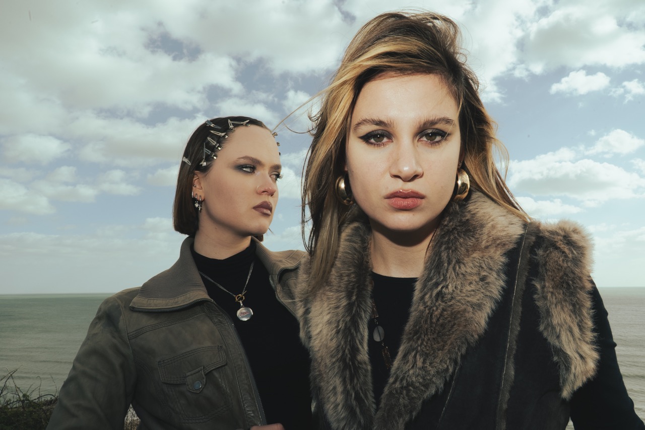 Two models pose outdoors near a coastline under a partly cloudy sky. The model on the left wears a grey jacket with hairclips, while the model on the right, in a fur vest, wears large earrings. The ocean is visible in the background.