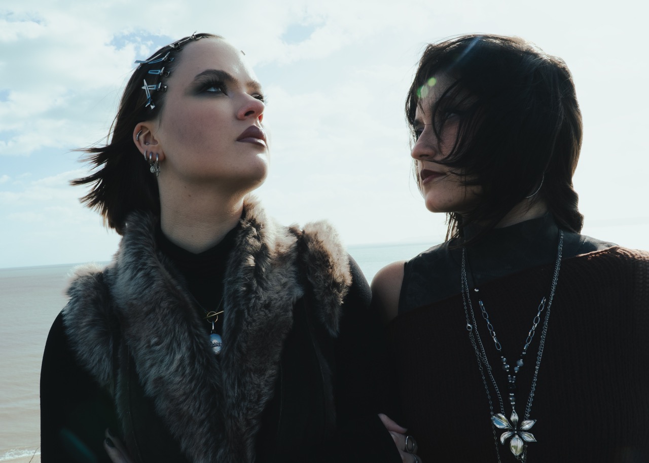 Two women standing together by the sea, both dressed in dark, stylish clothing. One woman wears a fur collar and has hair accessories, while the other has short hair and layered necklaces. The background features a cloudy sky and the ocean.