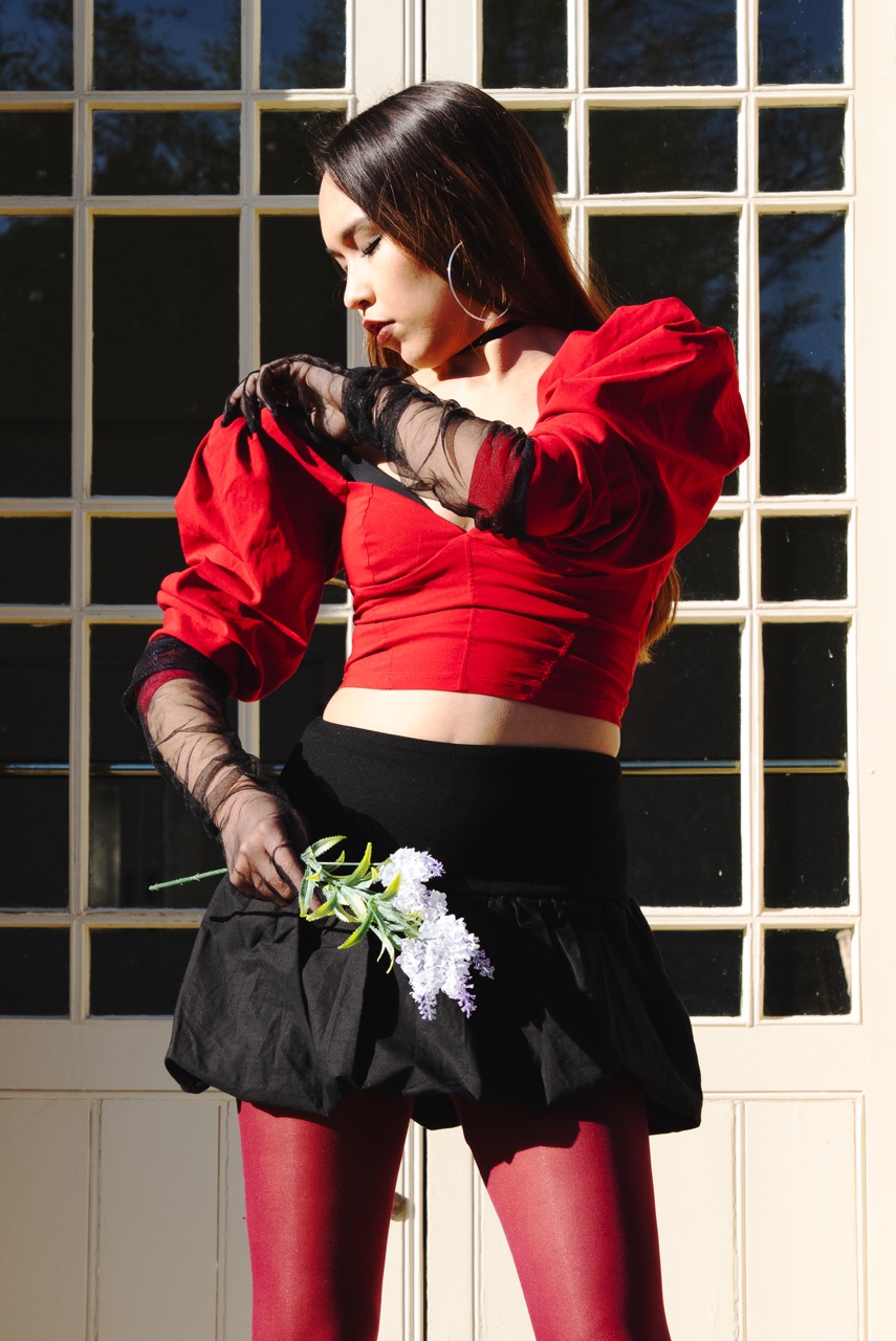 A woman wearing a red cropped top with puffed sleeves, a black skirt, and red tights stands in front of a large window. She holds a small bouquet of white flowers and is looking down at them.