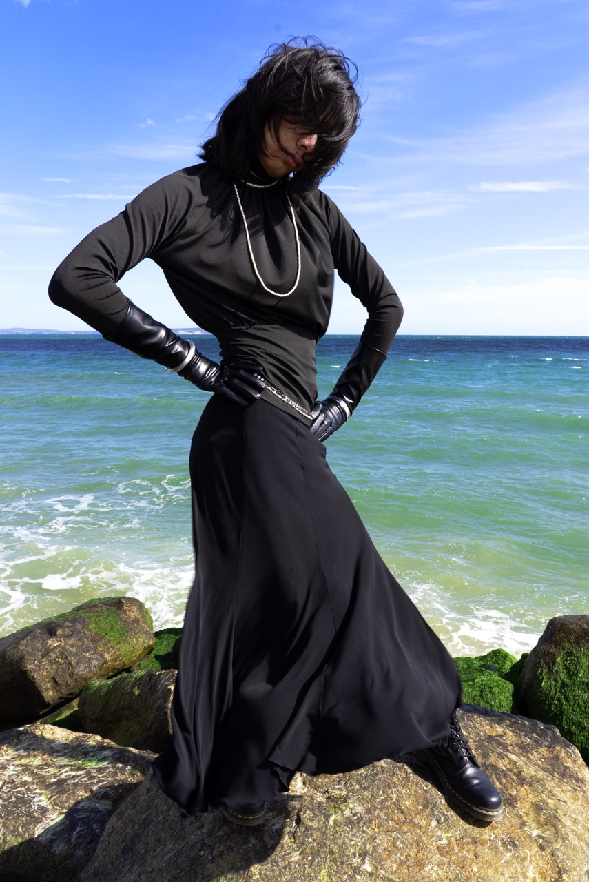 A person stands on a rocky shoreline wearing a long black skirt, a fitted black top, and black gloves. They pose with hands on hips against a backdrop of a blue sea and a clear sky.