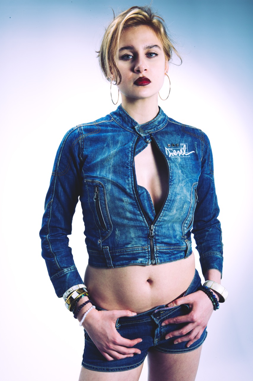 A young woman wearing a short denim jacket with a zip and matching denim shorts, posing with her hands on her hips. She has short blonde hair and large hoop earrings. The background features a gradient effect, transitioning from light to dark.
