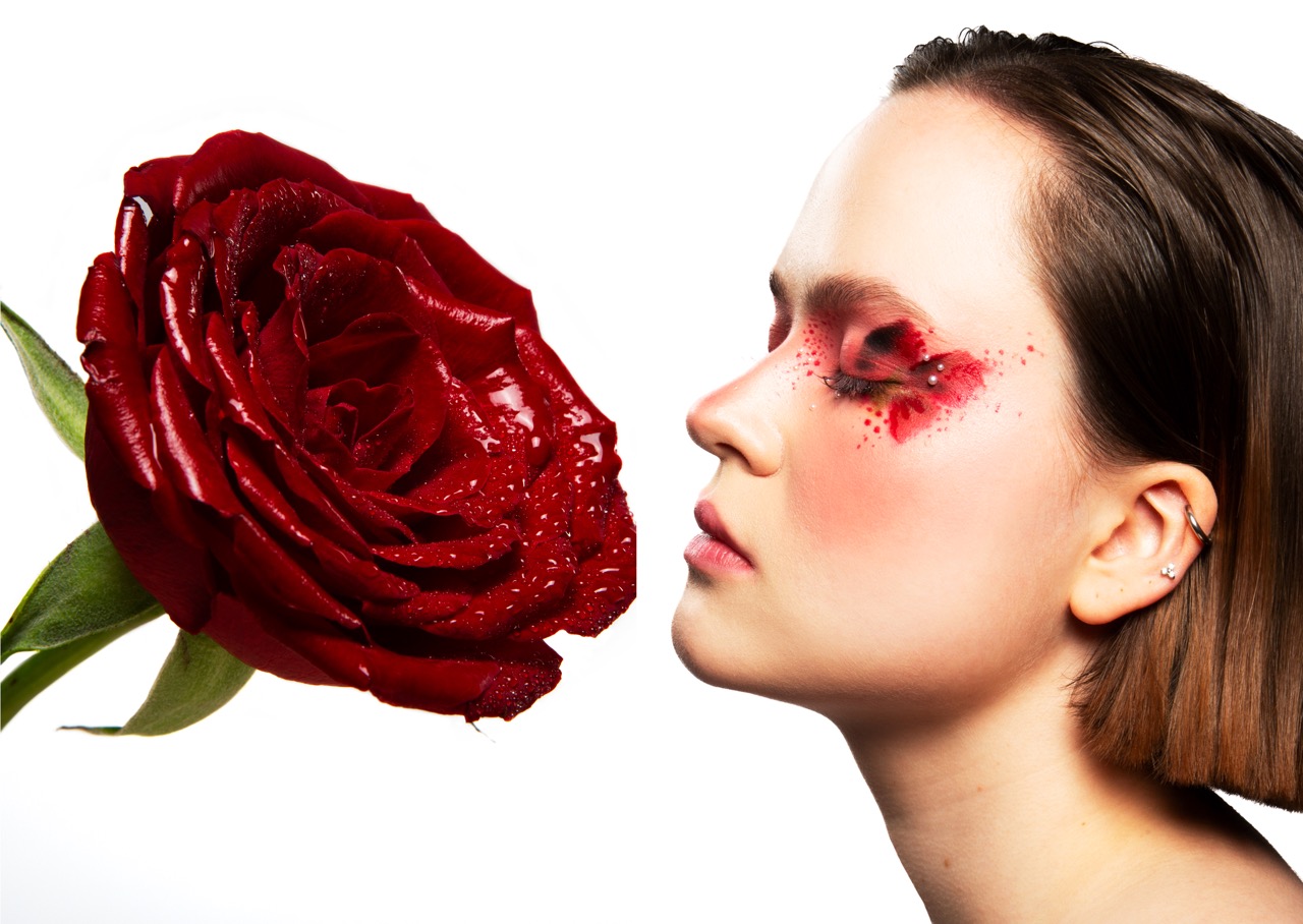 A close-up profile of a woman with styled hair, facing a large red rose. The woman's face features artistic makeup in shades of pink and red, with decorative dots around her eye. The background is a plain white, highlighting both the rose and her features.