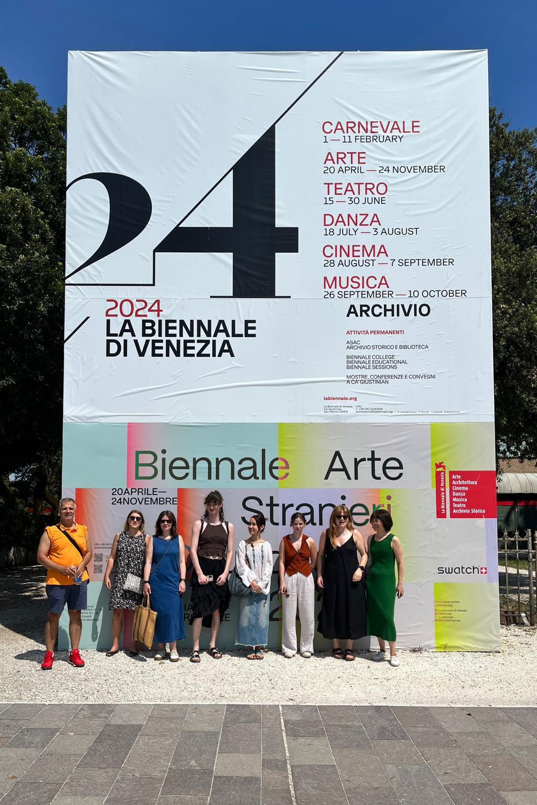 A group of people lined up in front of a poster for La Biennale di Venezia 2024 and posing for a photo.