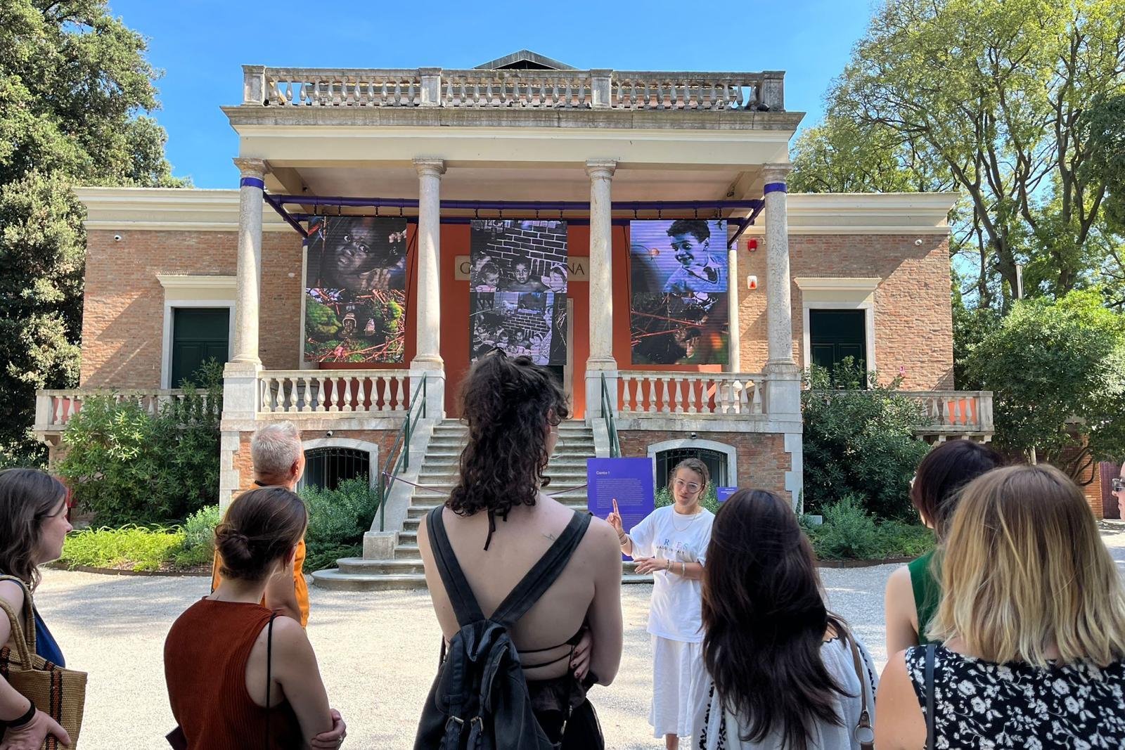 A group of people gathered in front of a pavilion in Venice. Three artworks appear to be on display in the pavilion.