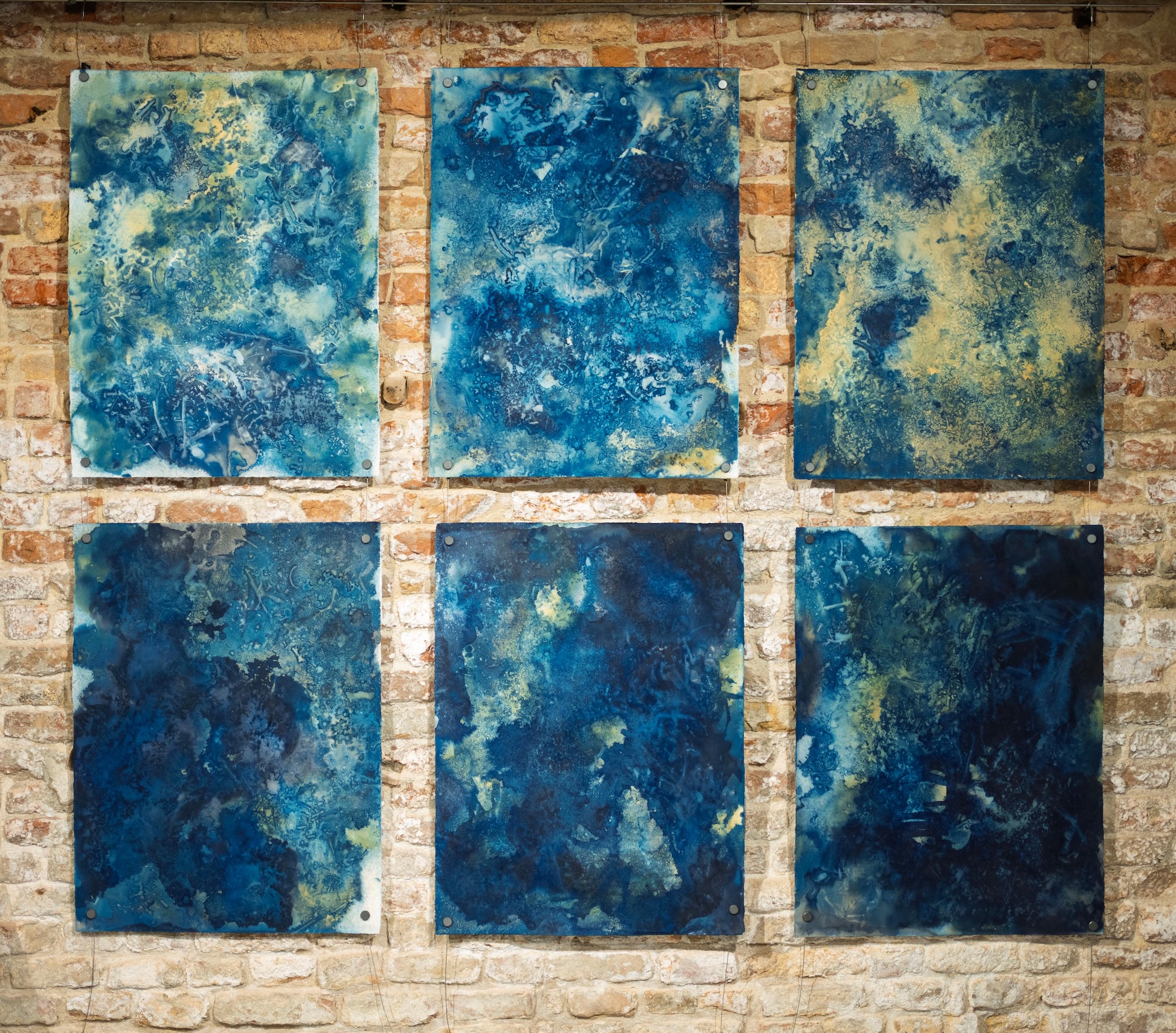 Six abstract blue-and-cream paintings arranged in a three-by-two grid on a brick wall.