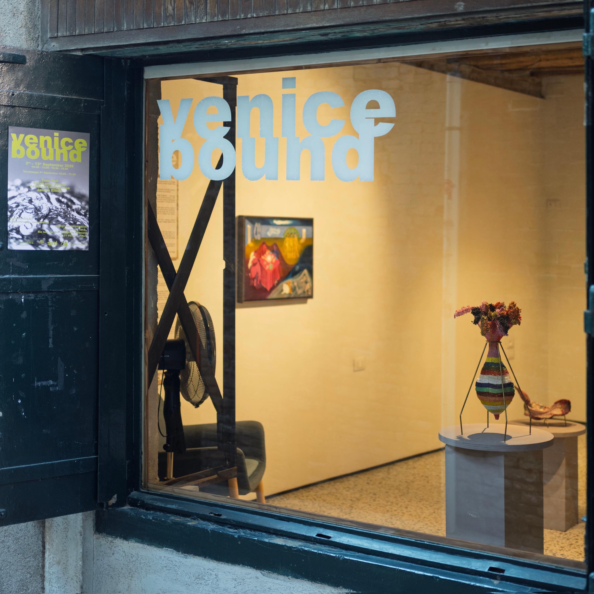 A gallery window displaying the Venice Bound title in pale blue lettering, with a framed painting and a striped vase on a pedestal inside.